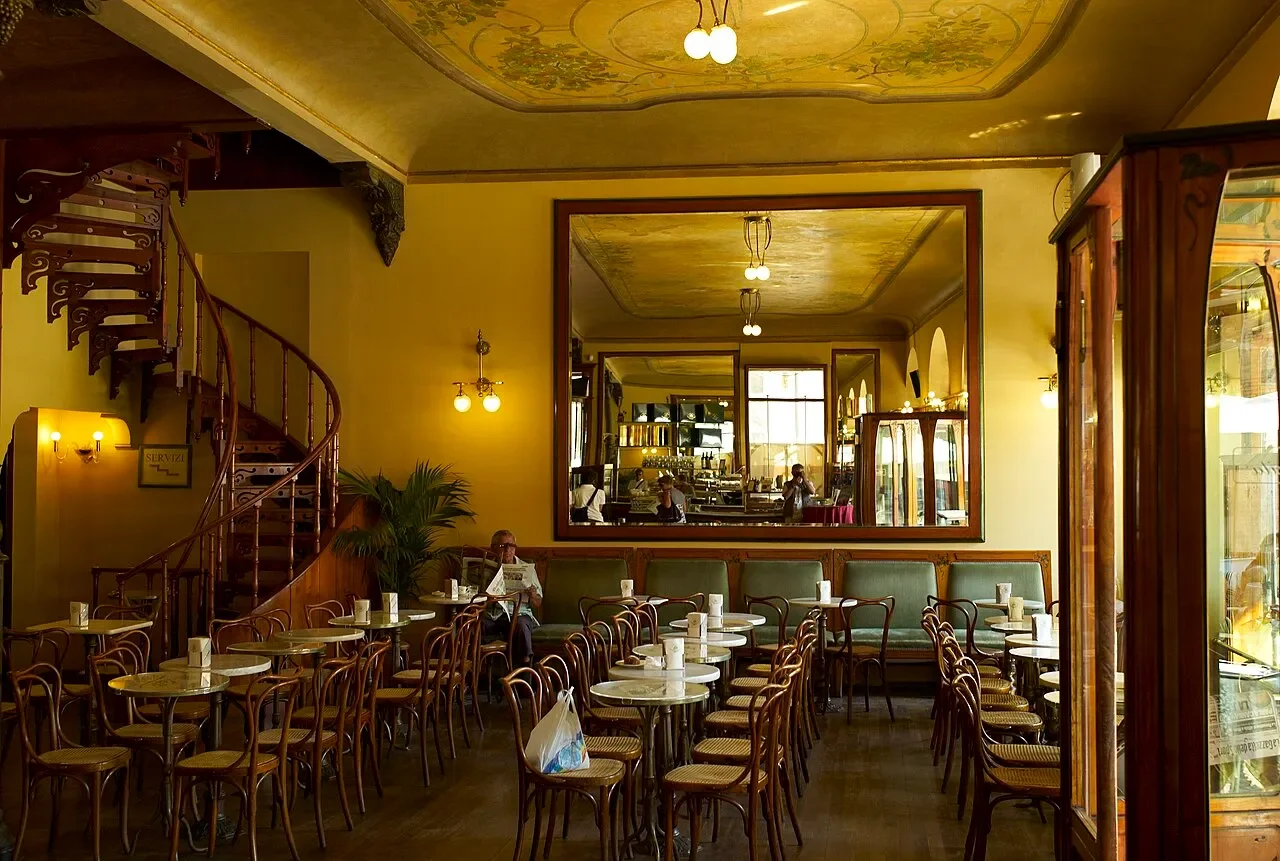 The historic interior of Caffè Meletti in Ascoli Piceno, with café tables, a spiral staircase and a painted ceiling