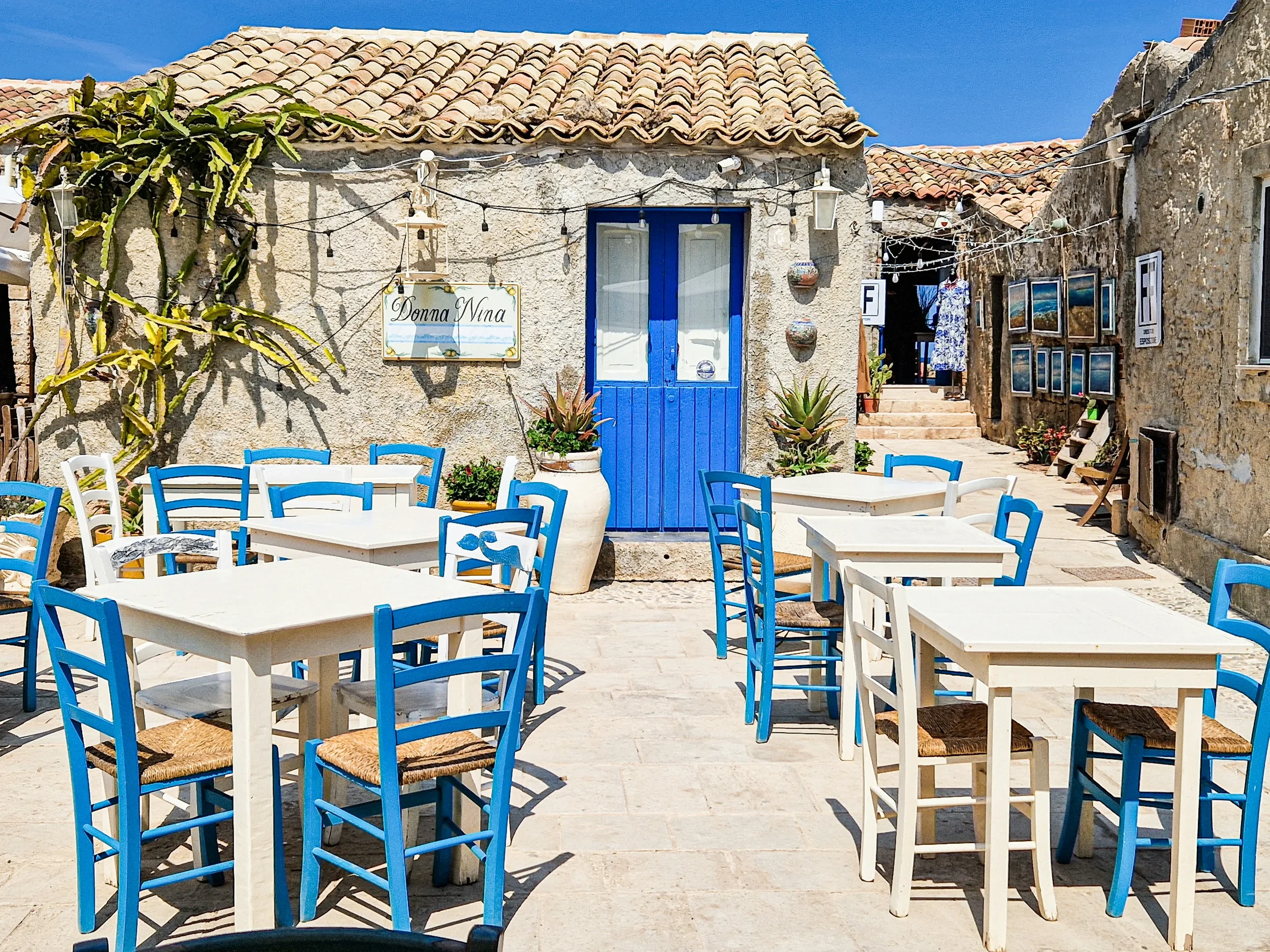 Blue wooden chairs and white restaurant tables outside a stone building with a blue door in Marzamemi