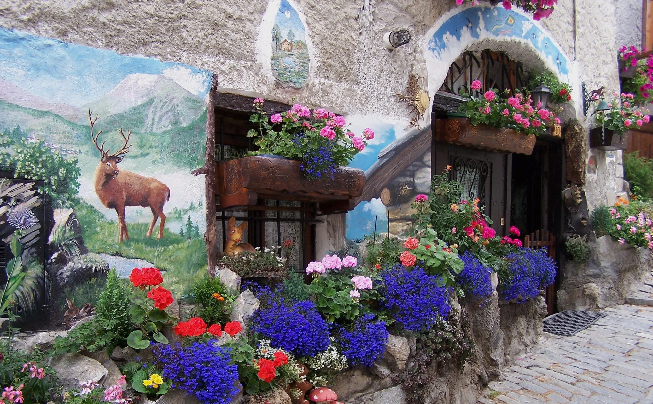 Flower-filled stone house in Usseaux, with a deer mural, mountain scenes and a painted arch beside a cobbled lane.
