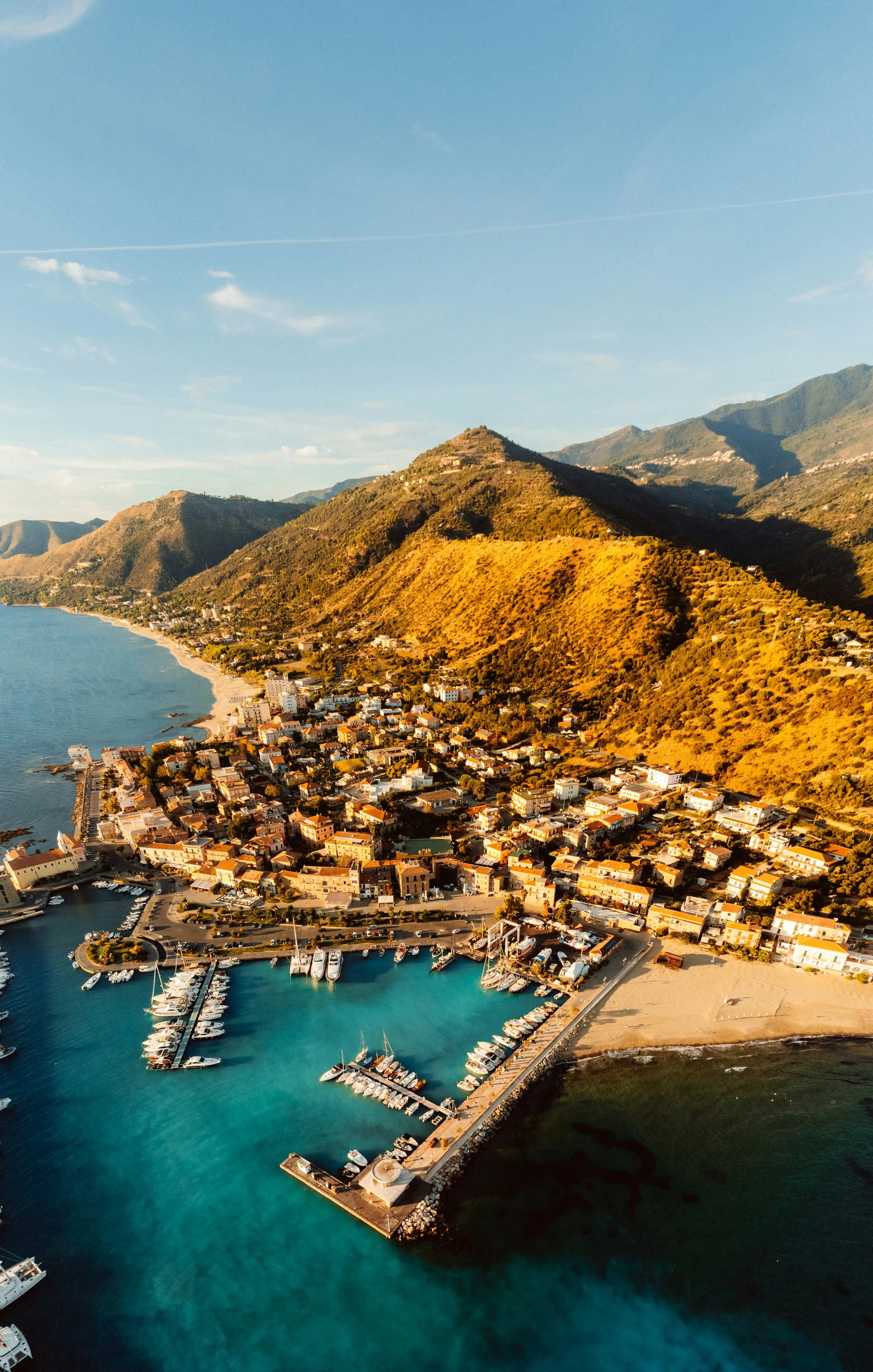 Aerial view of Acciaroli harbour, with moored boats, waterfront houses and hills behind the coast.