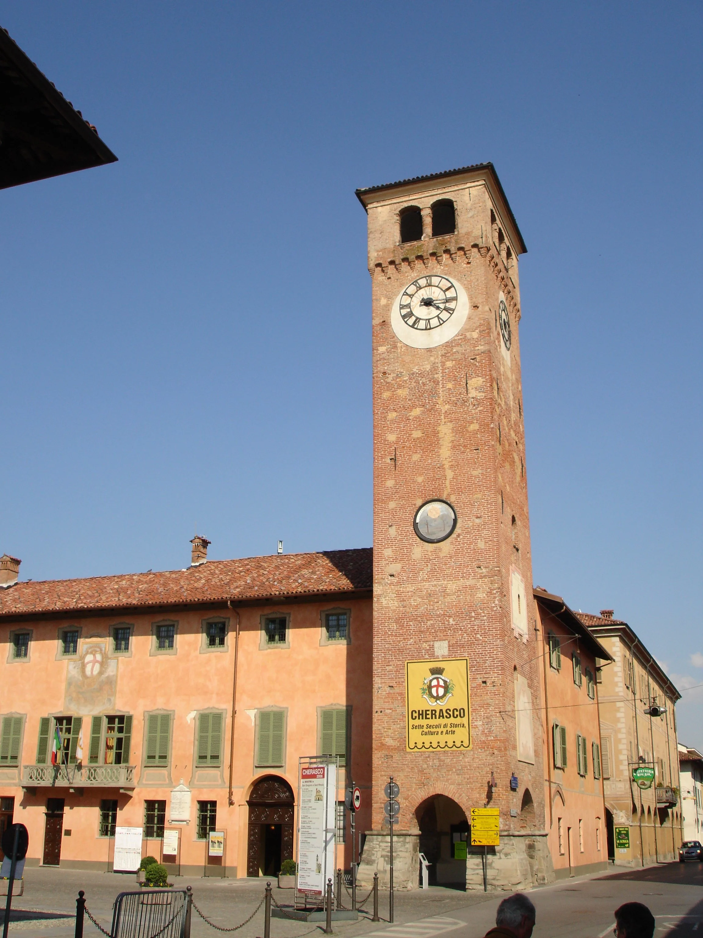 Cherasco’s brick civic clock tower beside the town hall, with green shutters and an arched entrance.