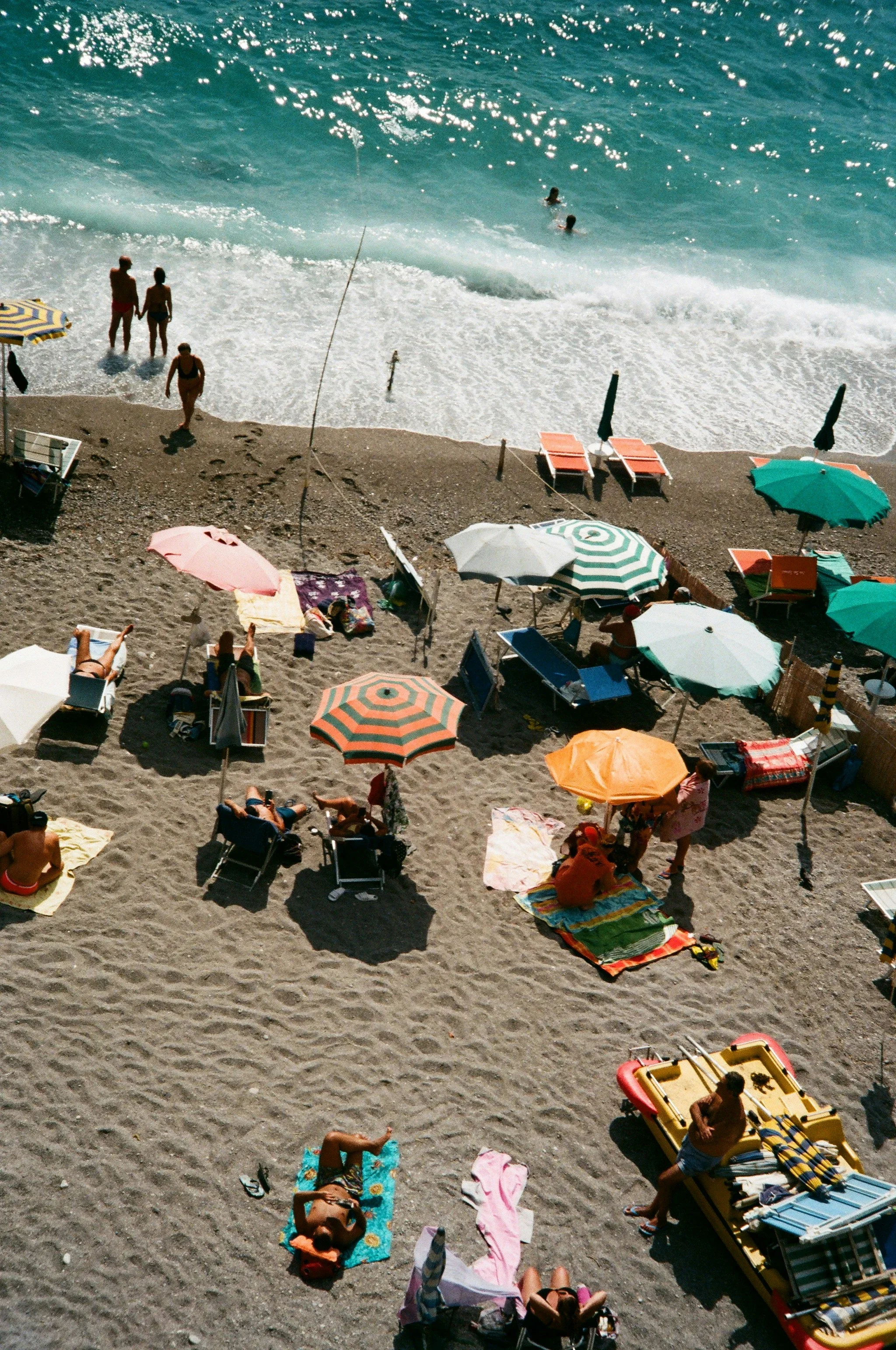 Colourful beach umbrellas and sun loungers beside turquoise waves on a dark sandy beach.