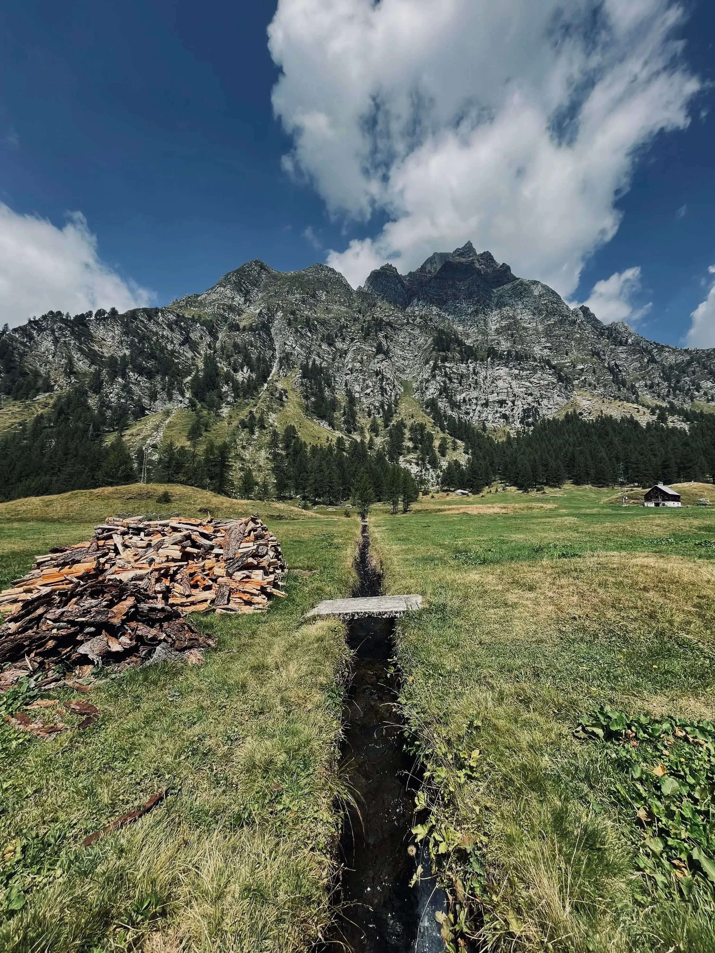 An Alpine meadow, a narrow stream and a stone hut beneath rocky mountain peaks.