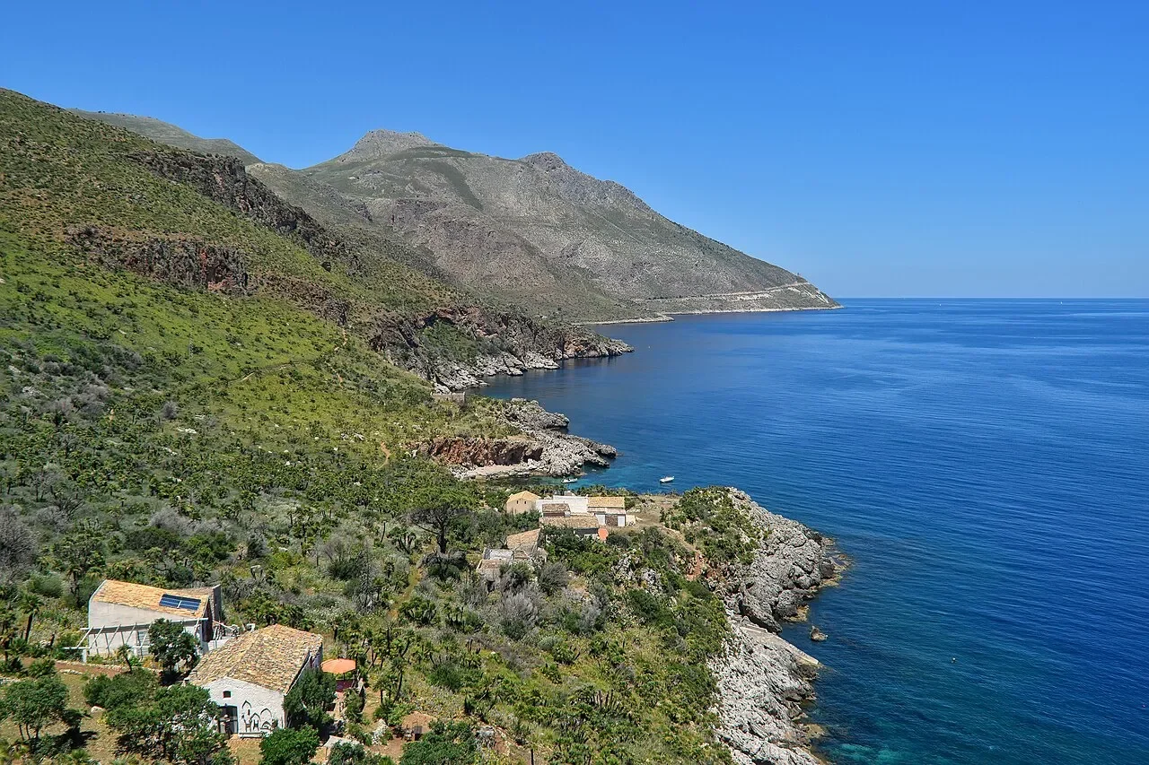 The rocky coastline of Zingaro Nature Reserve seen from the coastal trail, with green slopes and stone houses above the sea