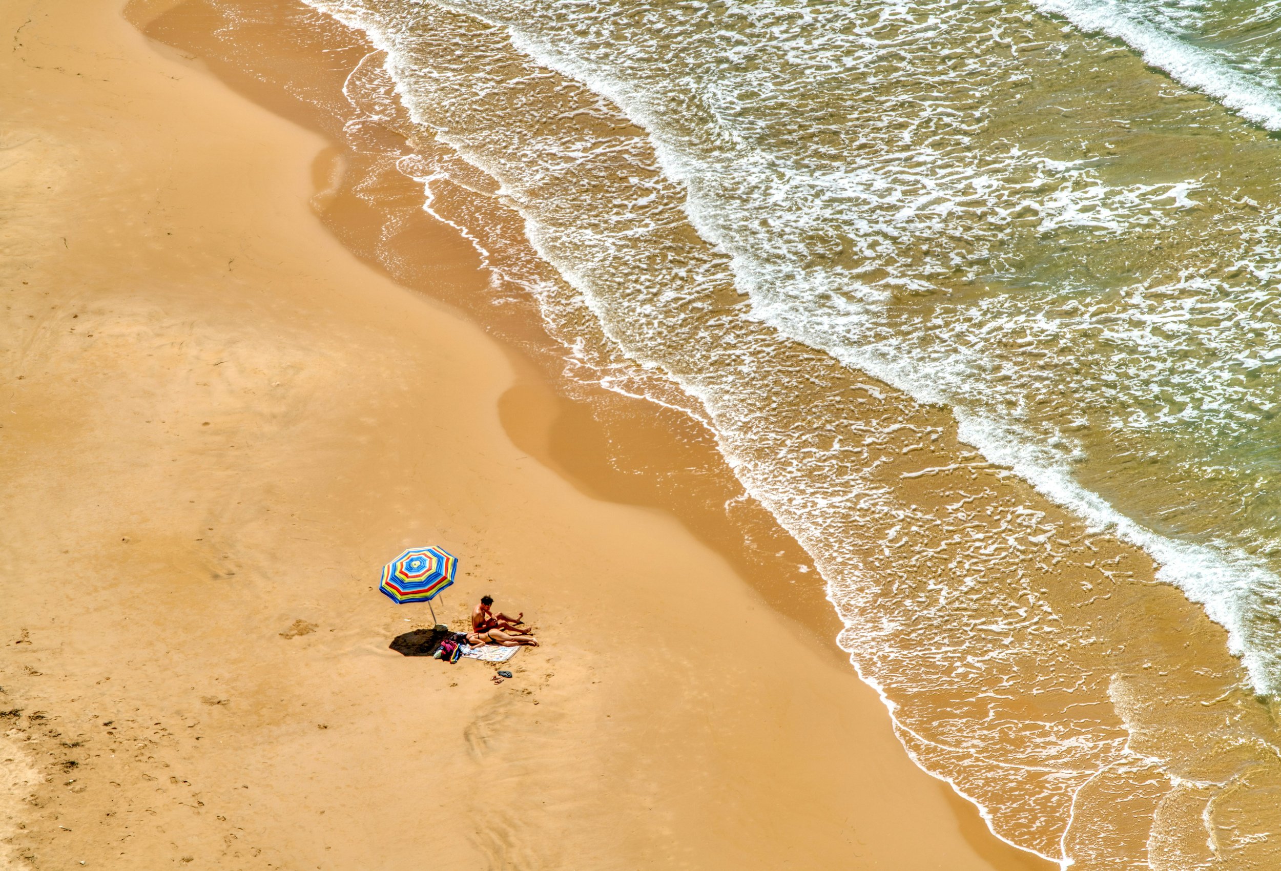 Aerial view of a golden sandy beach with turquoise waves and a colourful parasol in Sicily, Italy