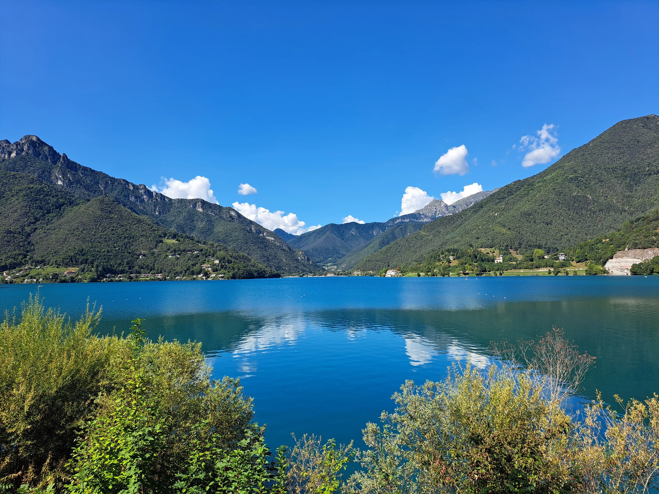 Blue lake water reflecting green mountains beneath a clear summer sky in Lombardia