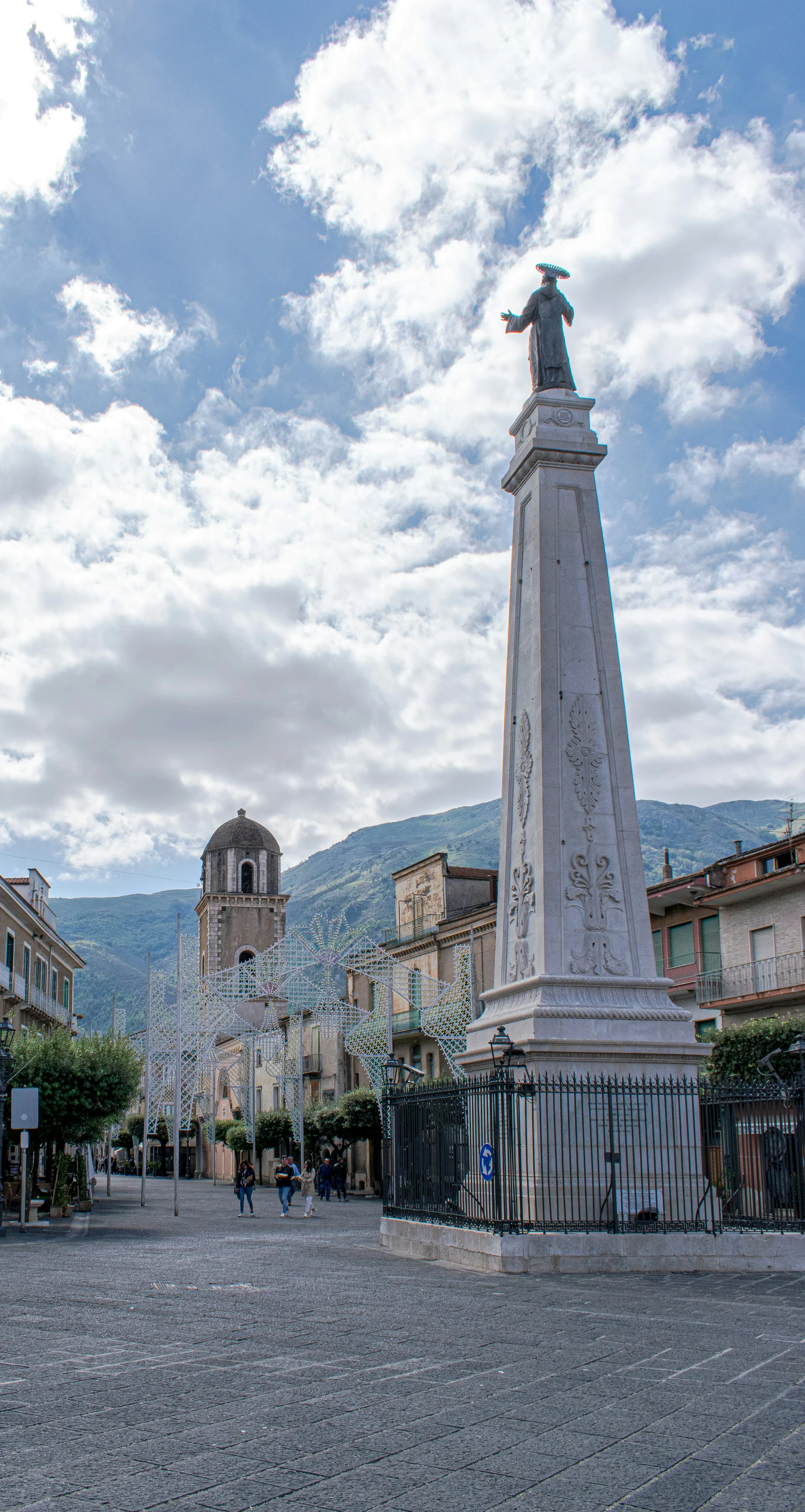 Monument topped by a saint’s statue in a Teggiano square, with a bell tower and mountains behind it.
