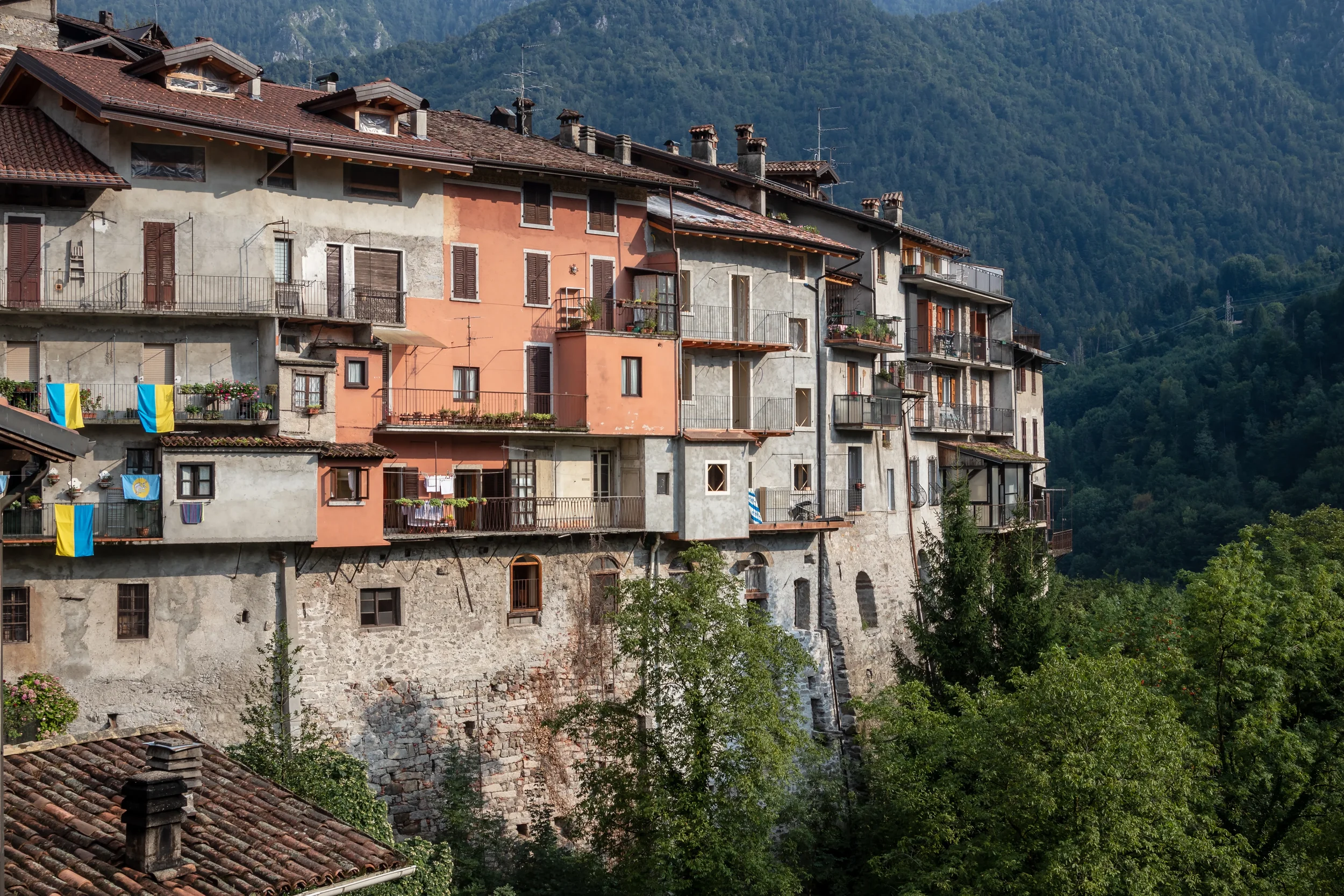 Traditional houses with balconies and tiled roofs in Bagolino, backed by a wooded mountain slope.