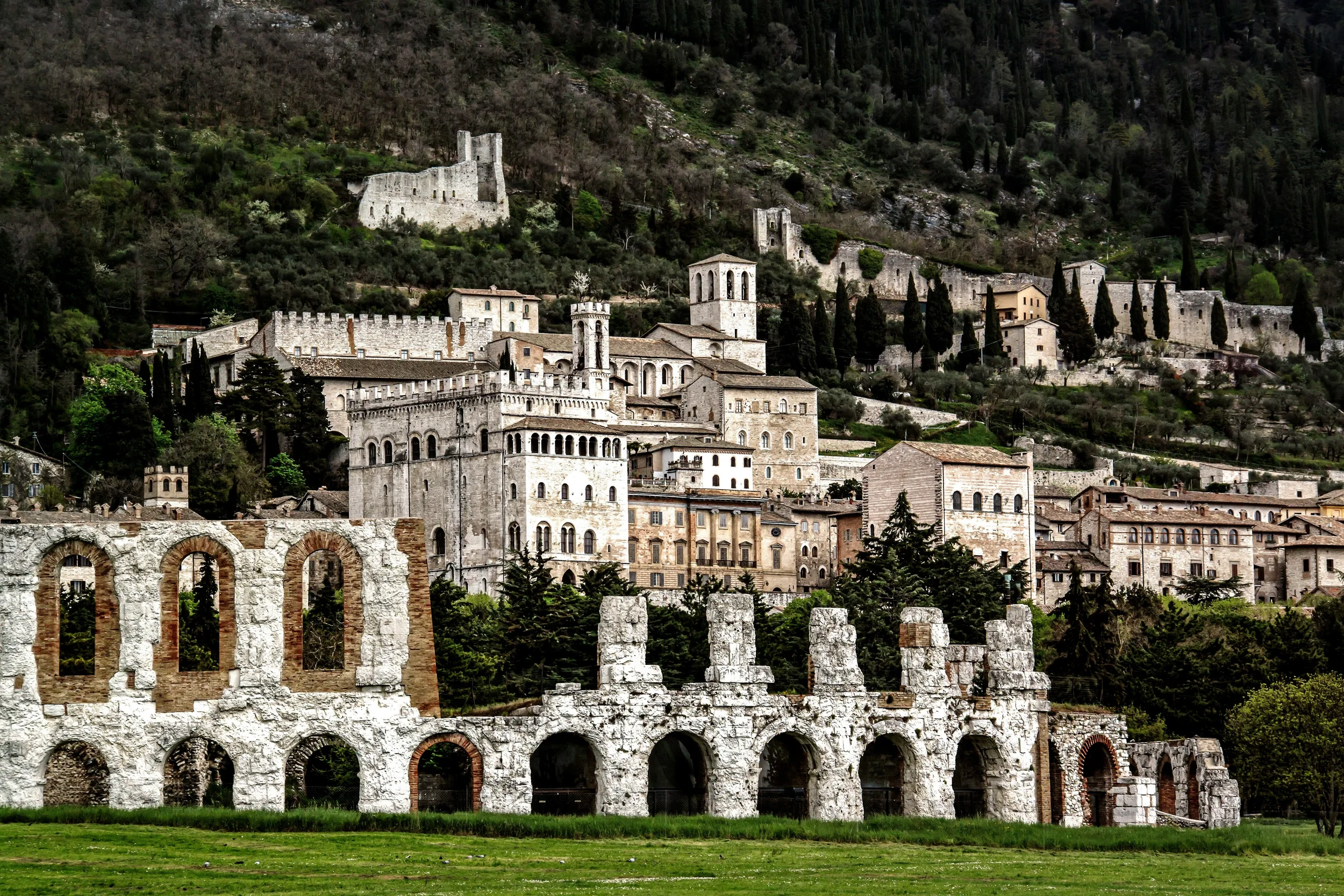 Roman theatre ruins in Gubbio, with the medieval town, stone towers and hillside fortifications in the background.