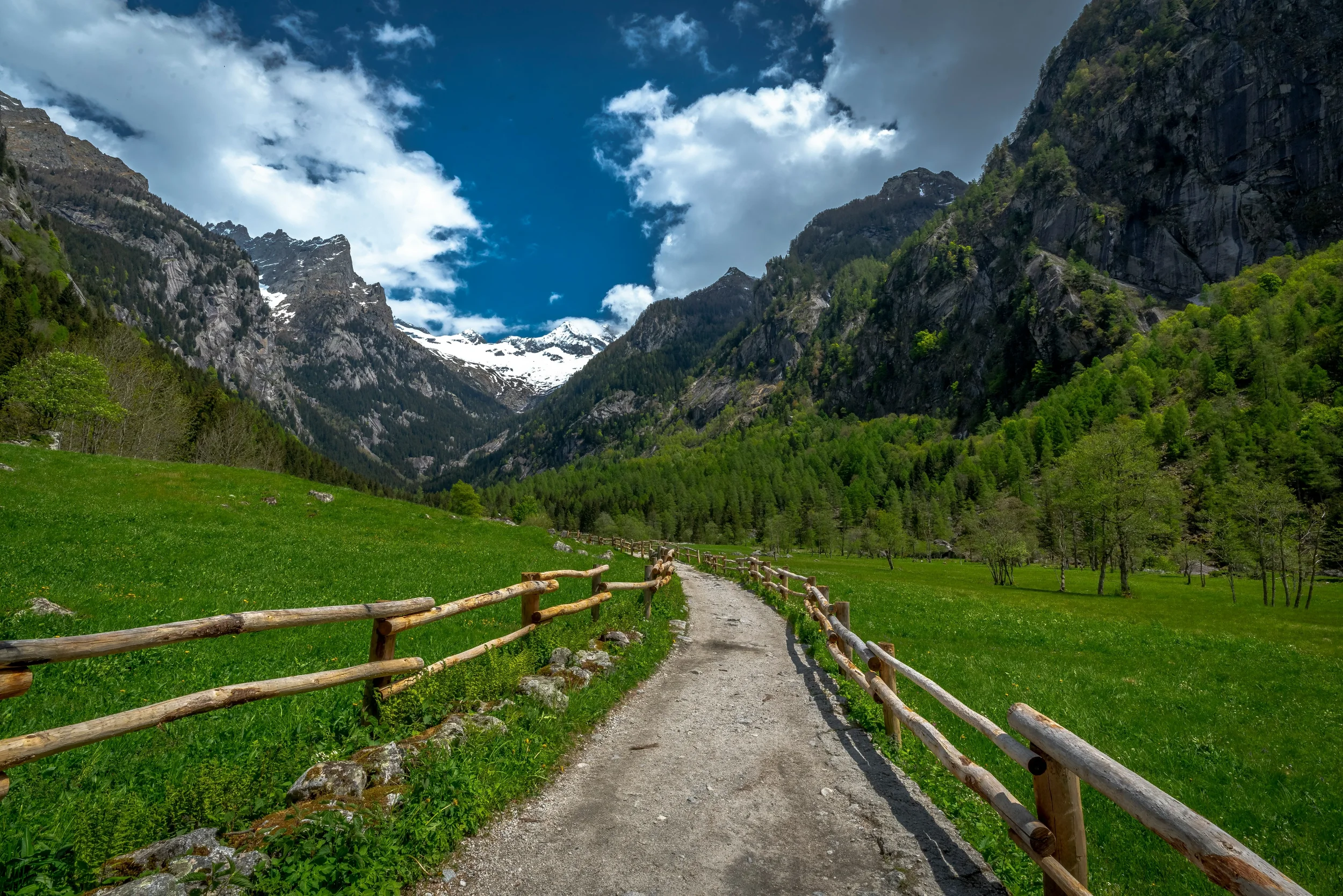 A fenced walking path through green meadows beneath granite cliffs and snowy peaks in Val di Mello.