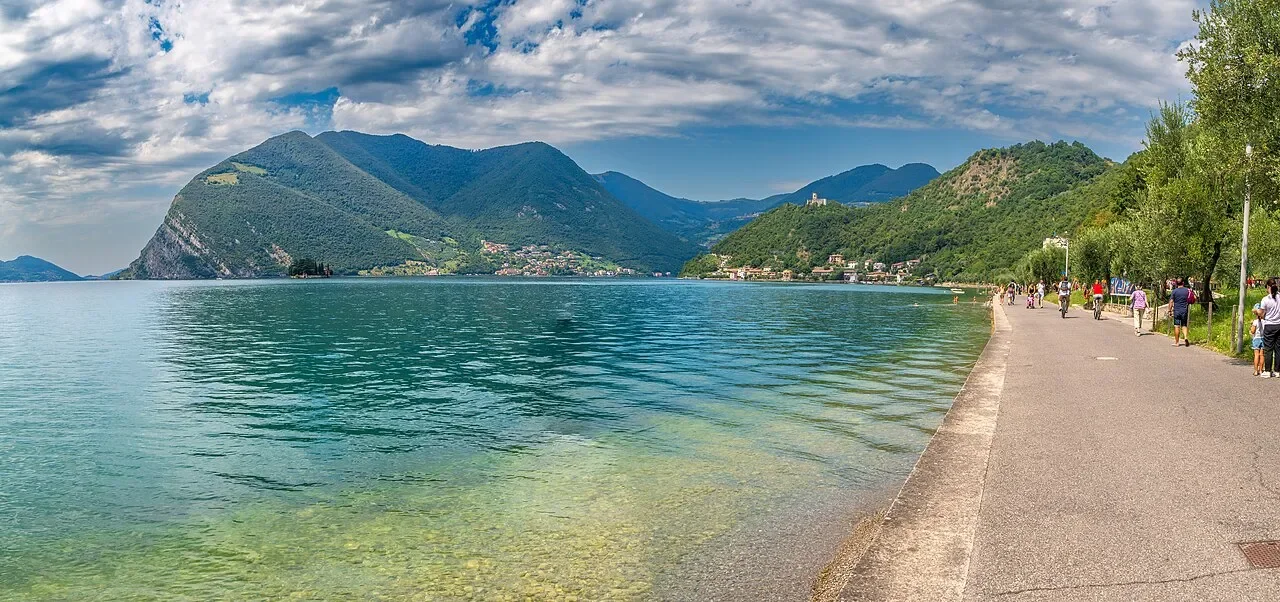 Walkers and cyclists on Monte Isola’s lakeside path beside clear water, with wooded mountains across Lake Iseo.