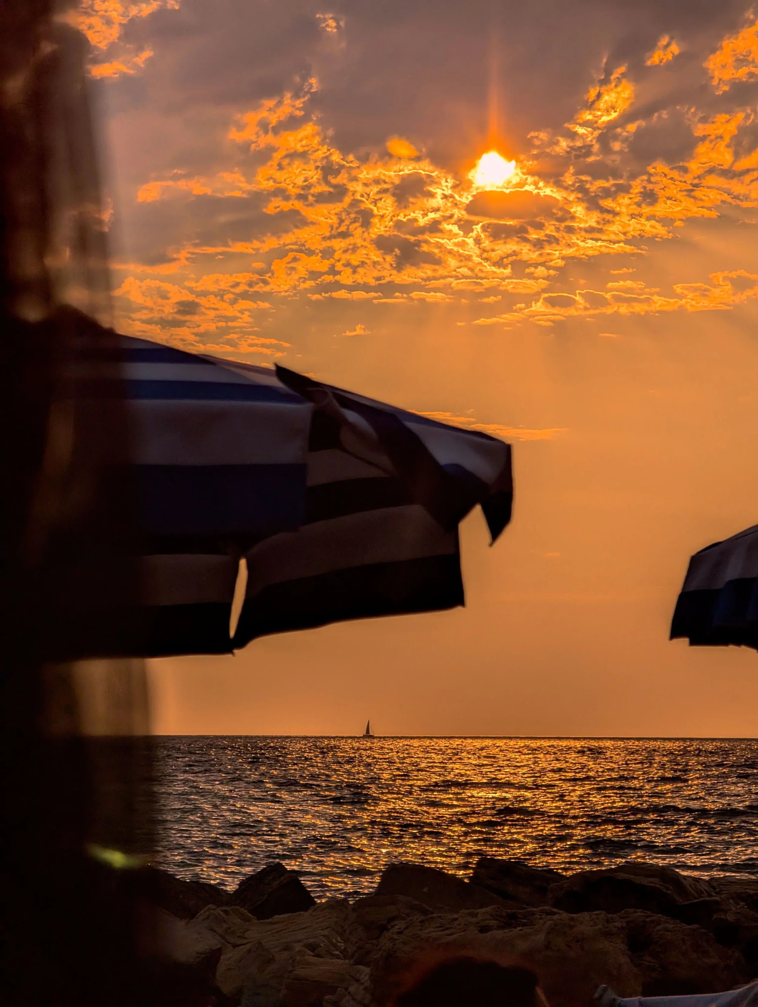 Golden sunset over the sea, with beach umbrellas and rocks along the shoreline.