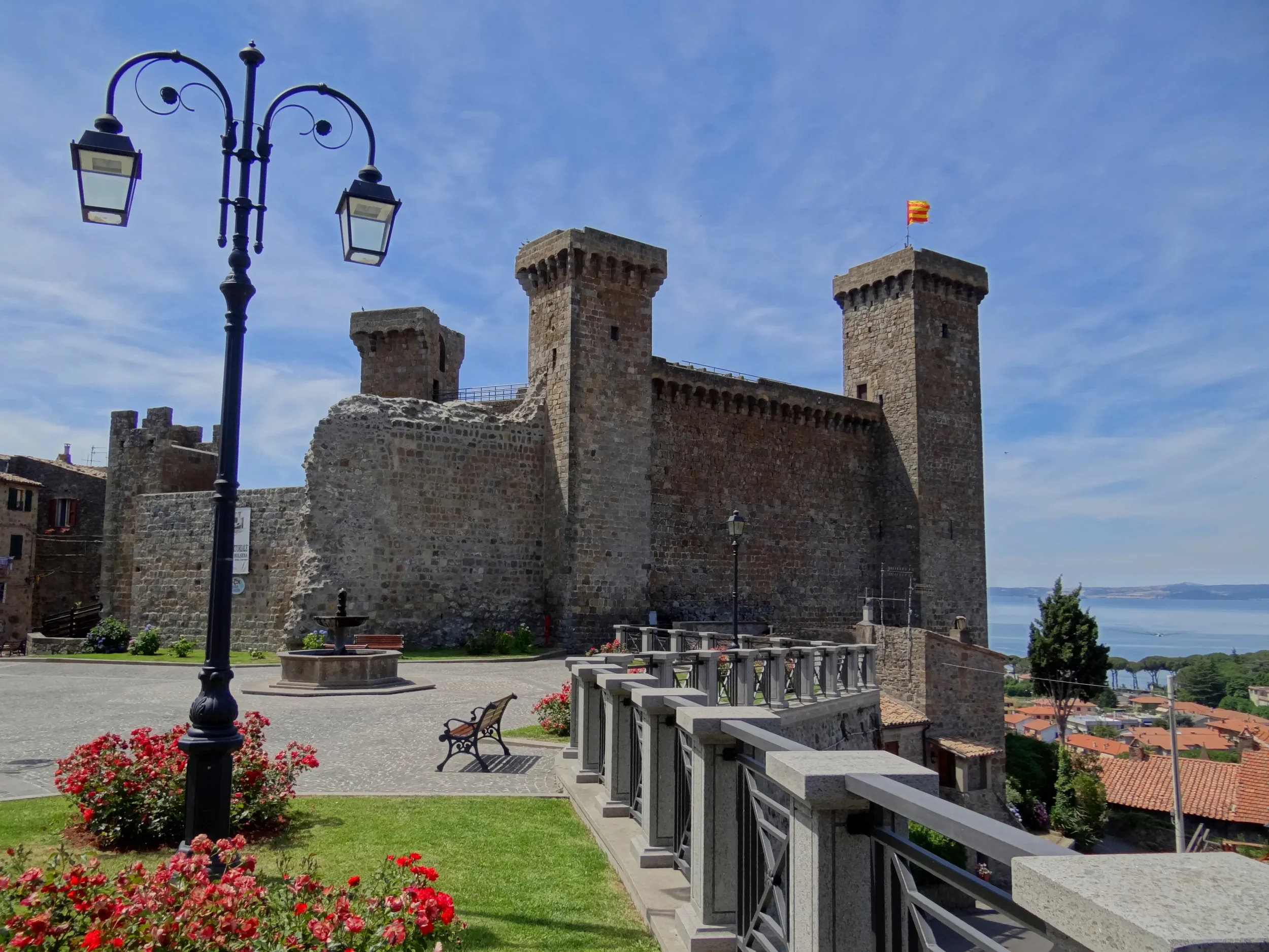 The stone towers of Rocca Monaldeschi in Bolsena, with flowers and Lake Bolsena beyond.