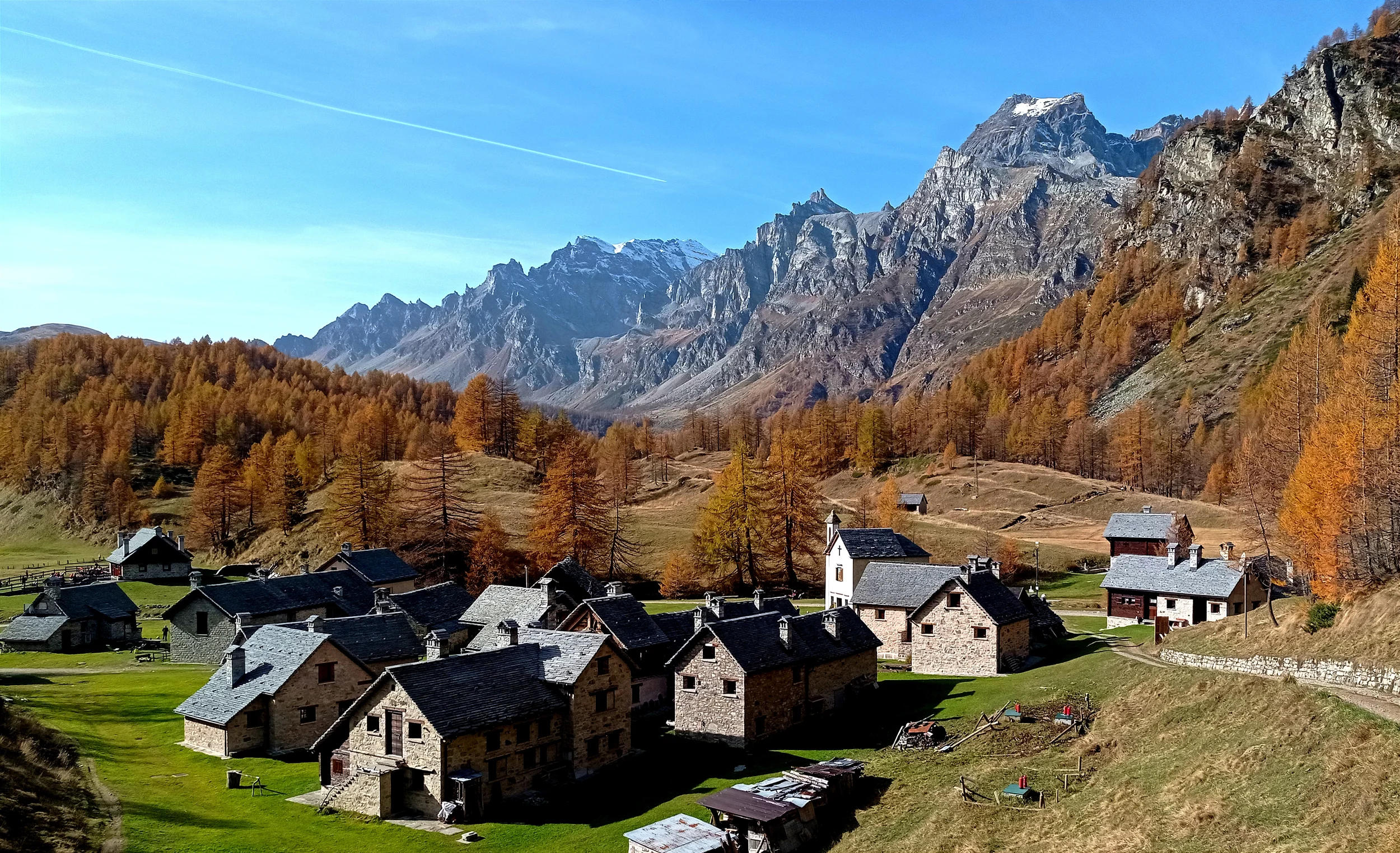 Stone-roofed houses and the small church of Crampiolo beneath golden larches and mountain peaks.