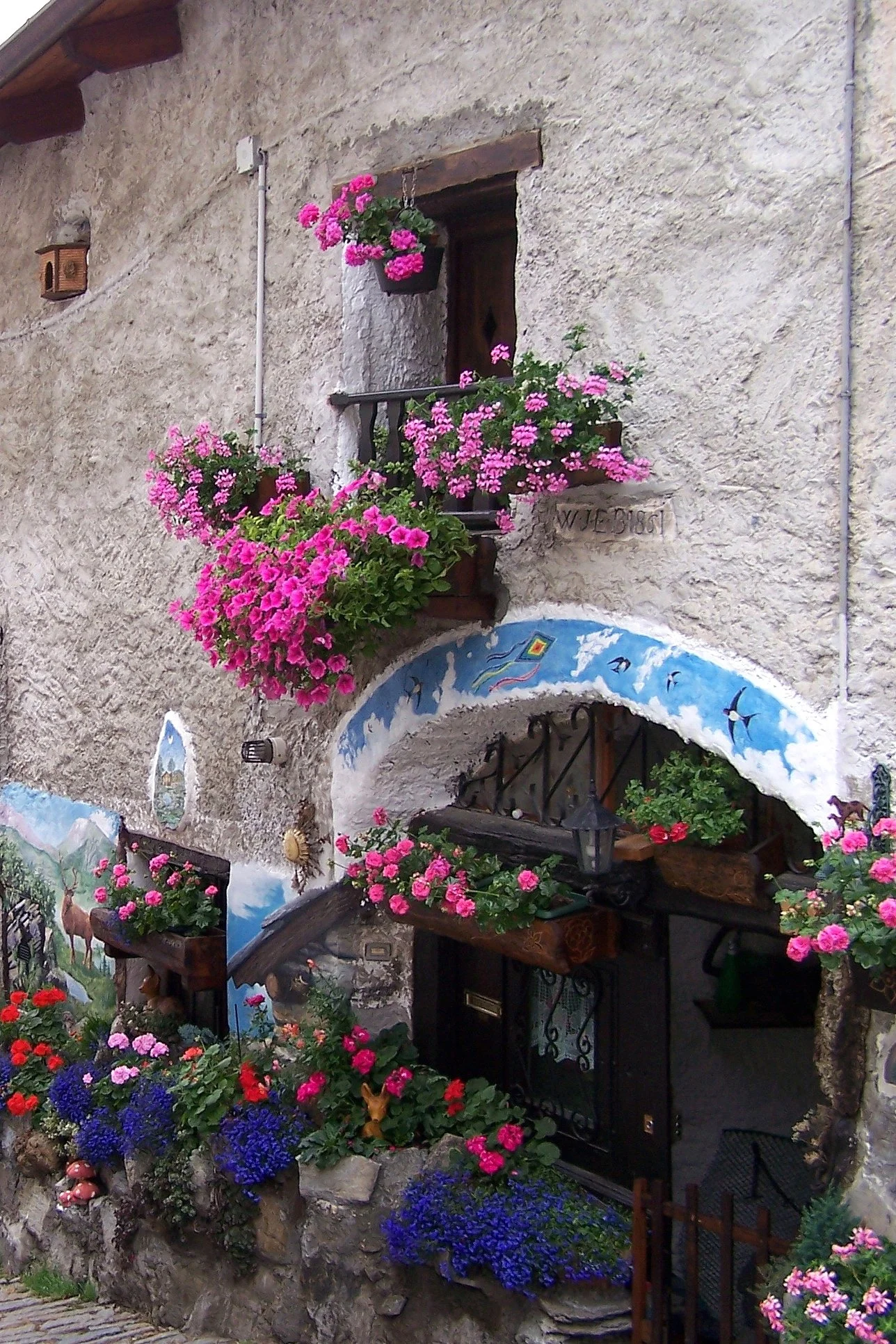 Pink and purple flowers around a painted archway and balcony on a stone house in Usseaux.