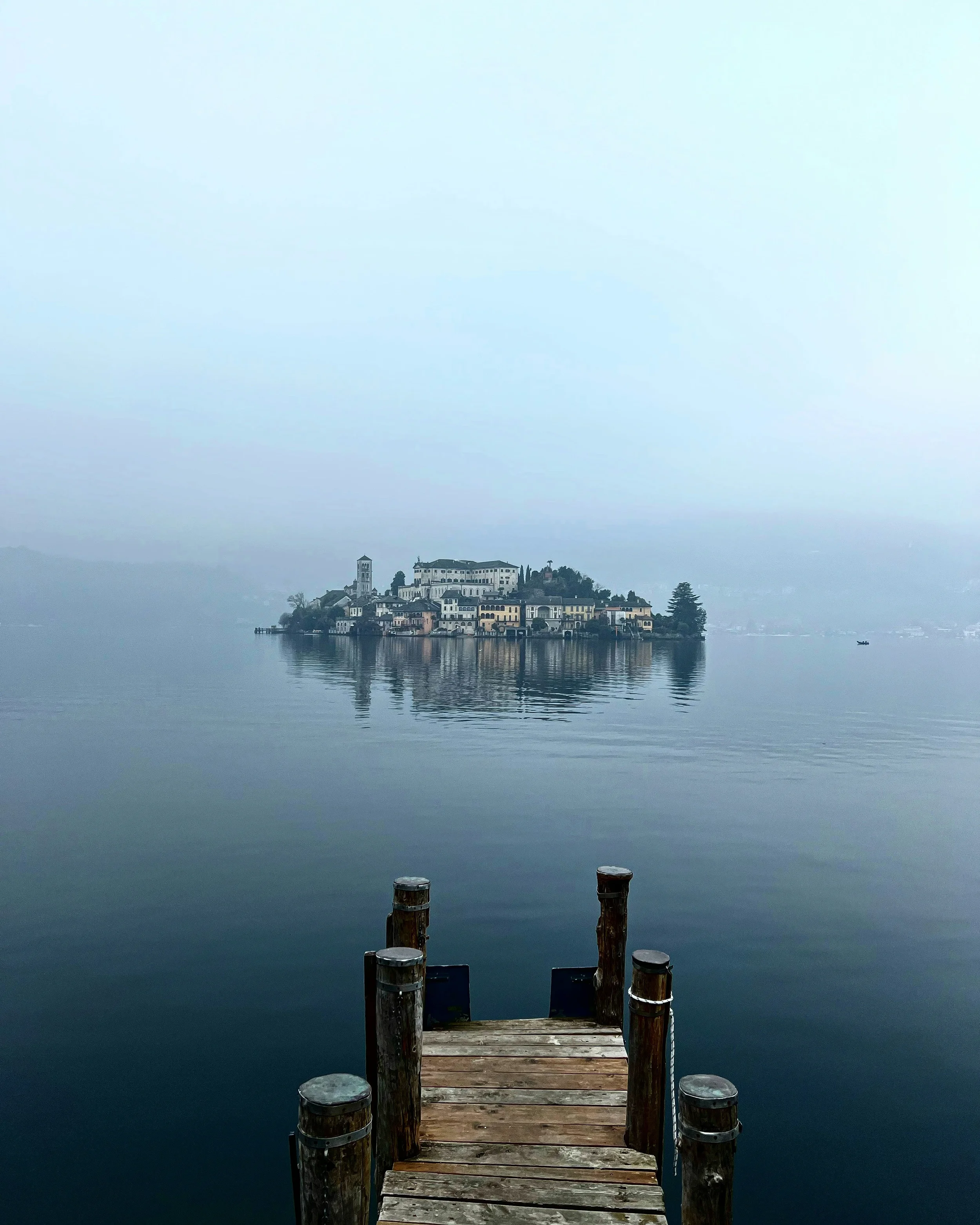 San Giulio Island reflected in misty Lake Orta, beyond a wooden jetty.