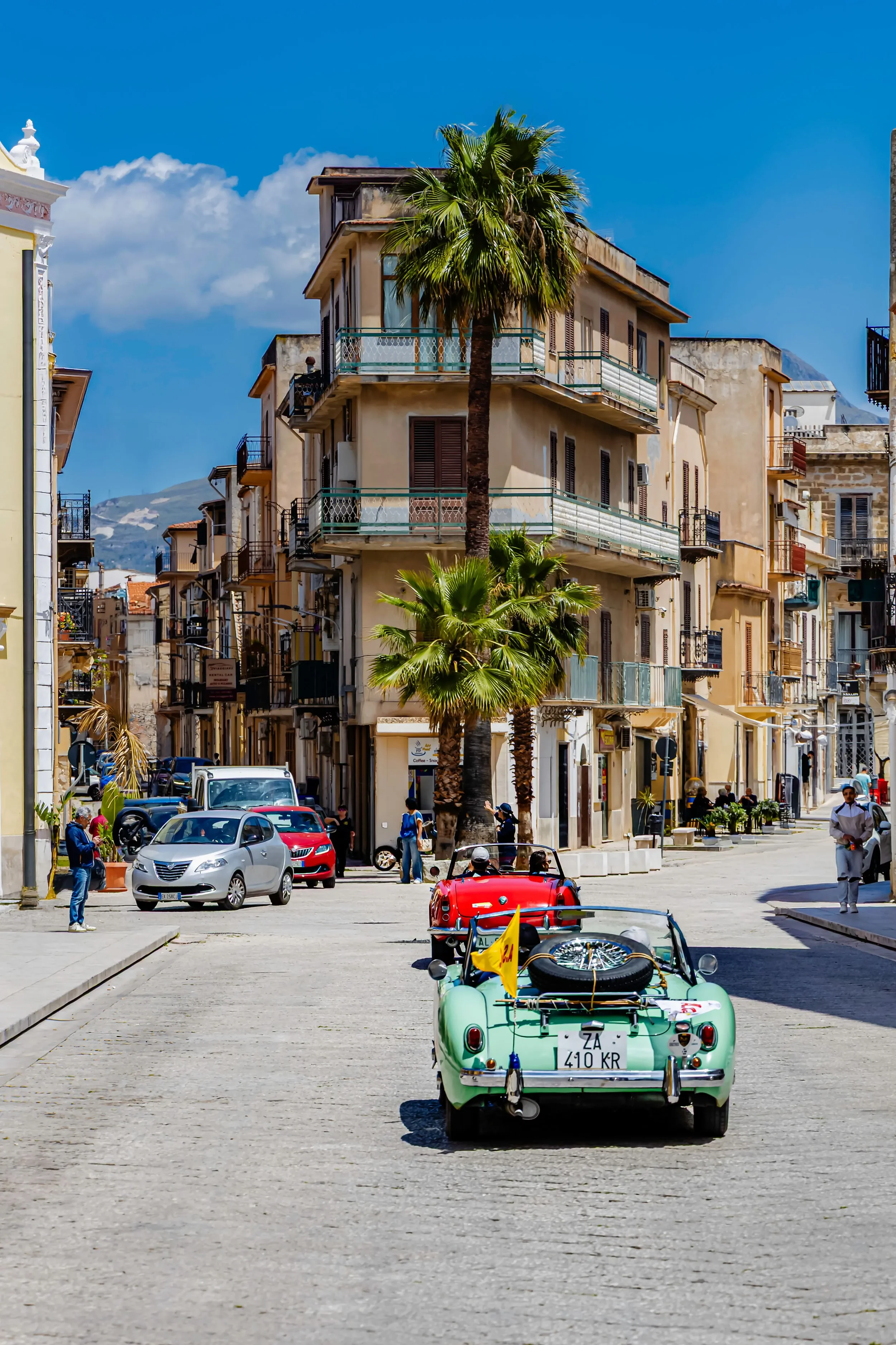 Palm-lined city street in Sicily with colourful vintage cars, historic buildings and lively traffic