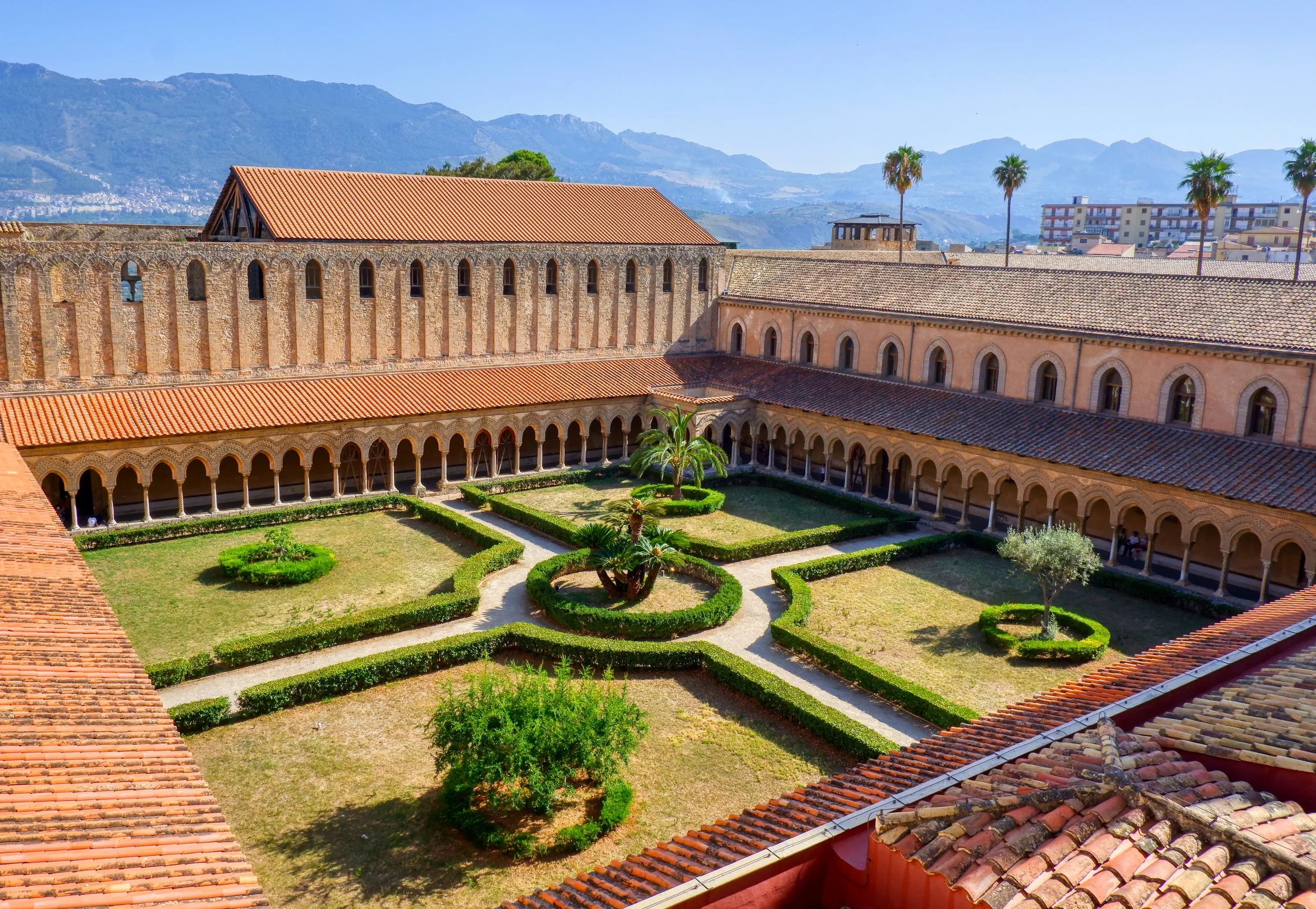 Monreale’s Benedictine cloister seen from above, with arcaded walkways surrounding a formal garden and mountains beyond