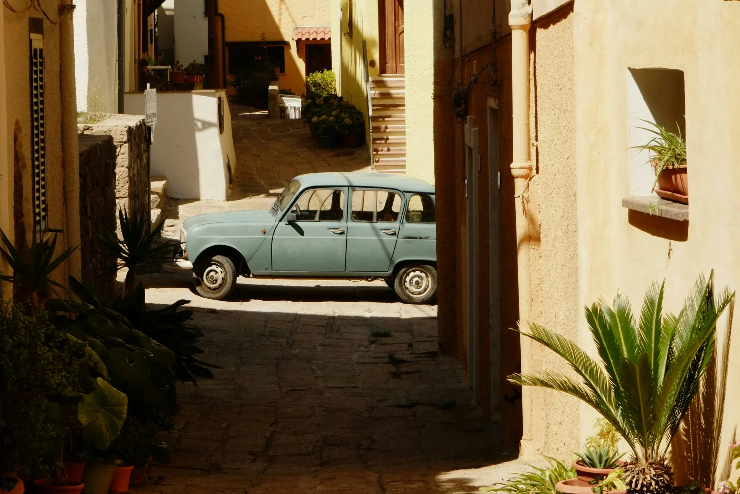 Vintage pale green car parked in a narrow village street in Sardinia, framed by traditional buildings and palm plants
