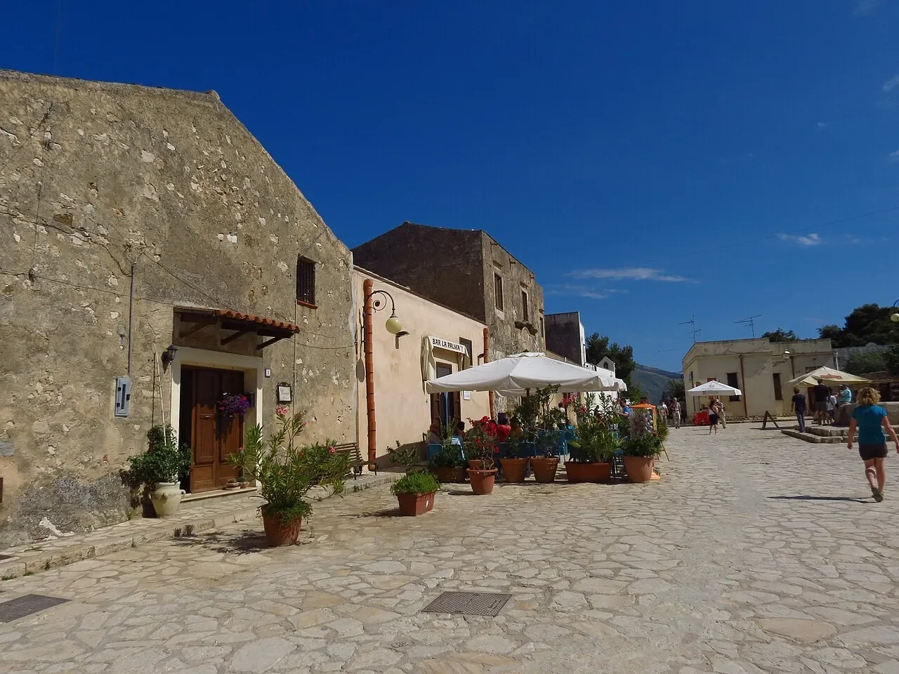 Stone buildings and café umbrellas around a sunlit paved square in Scopello village