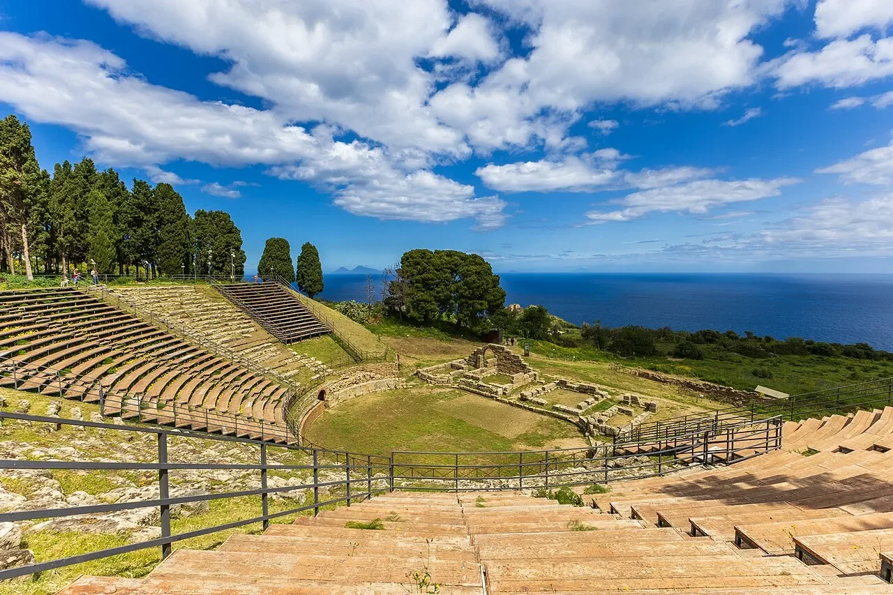 Curved stone seating in Tindari’s ancient theatre, with trees and the Tyrrhenian Sea beyond