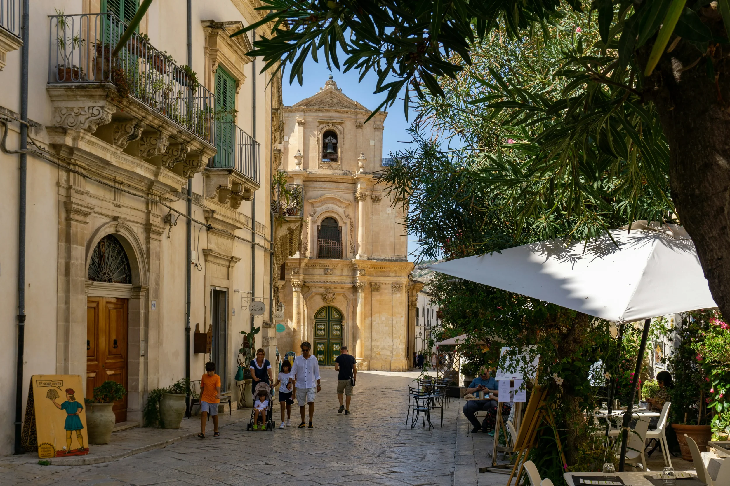 A pedestrian street in Scicli lined with carved stone balconies, café tables and a Baroque church