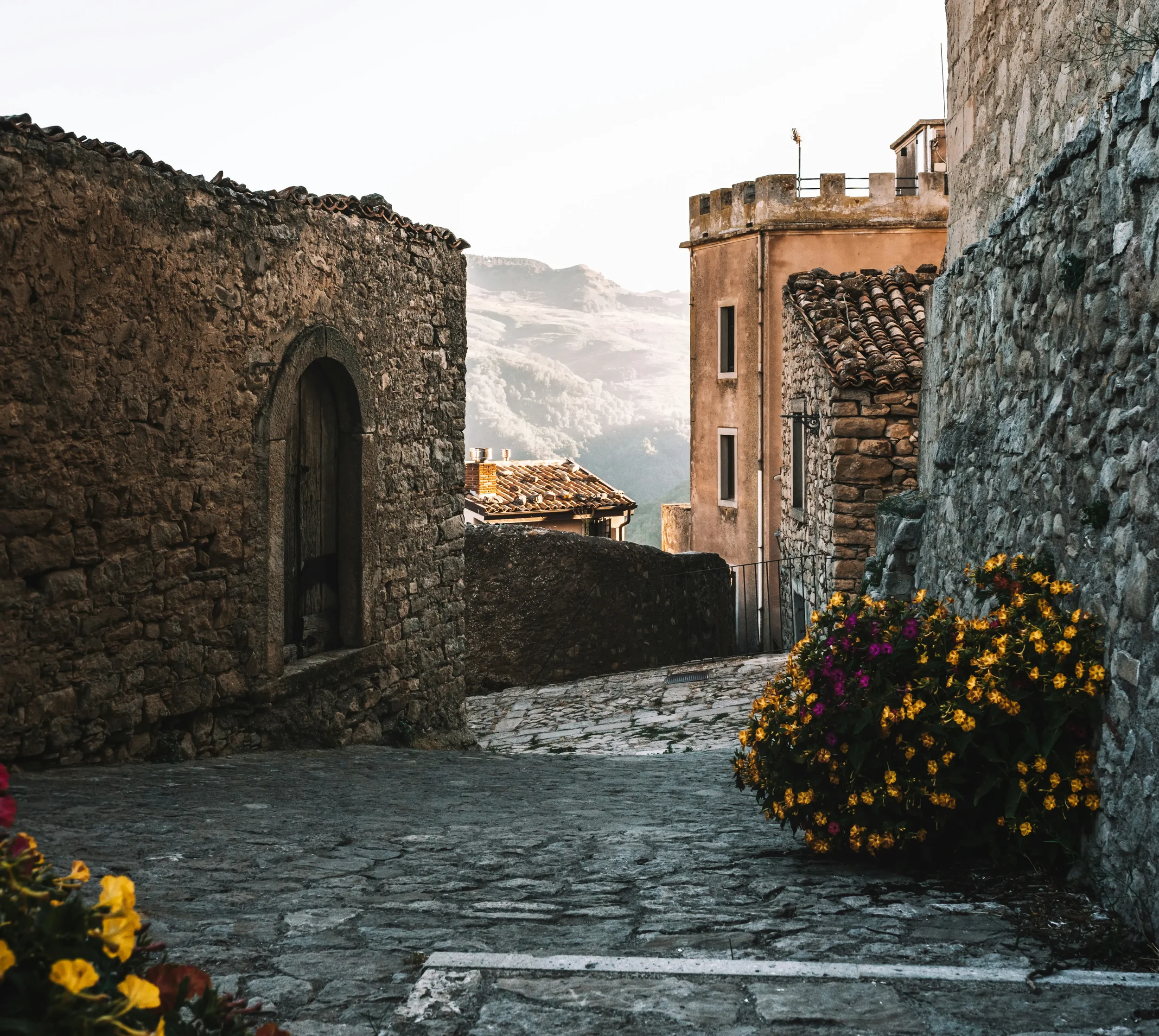 A cobbled lane between stone houses in Montalbano Elicona, with flowers beside the wall and hills beyond