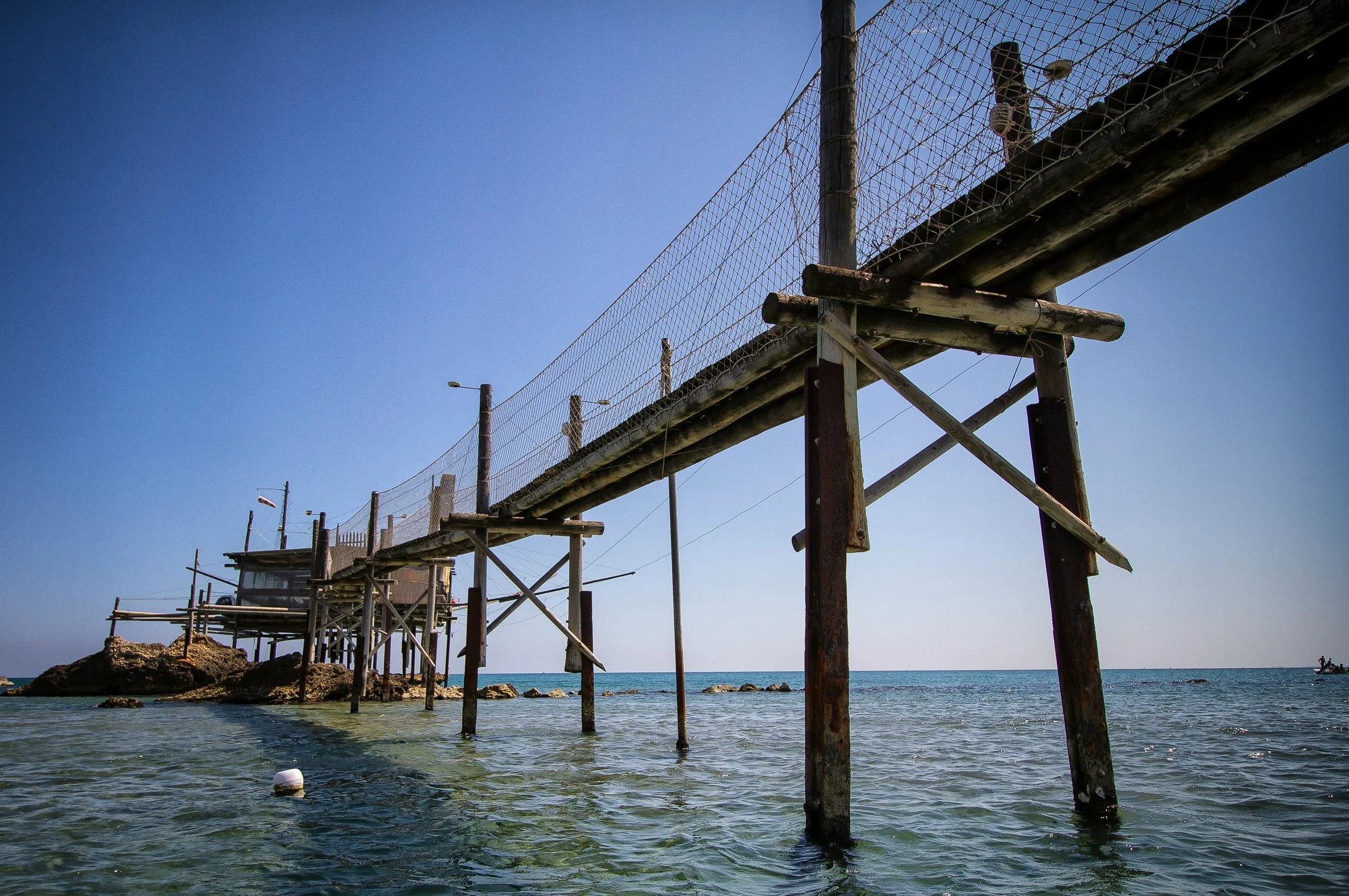 Wooden walkway leading to a traditional trabocco fishing platform above clear blue seawater.