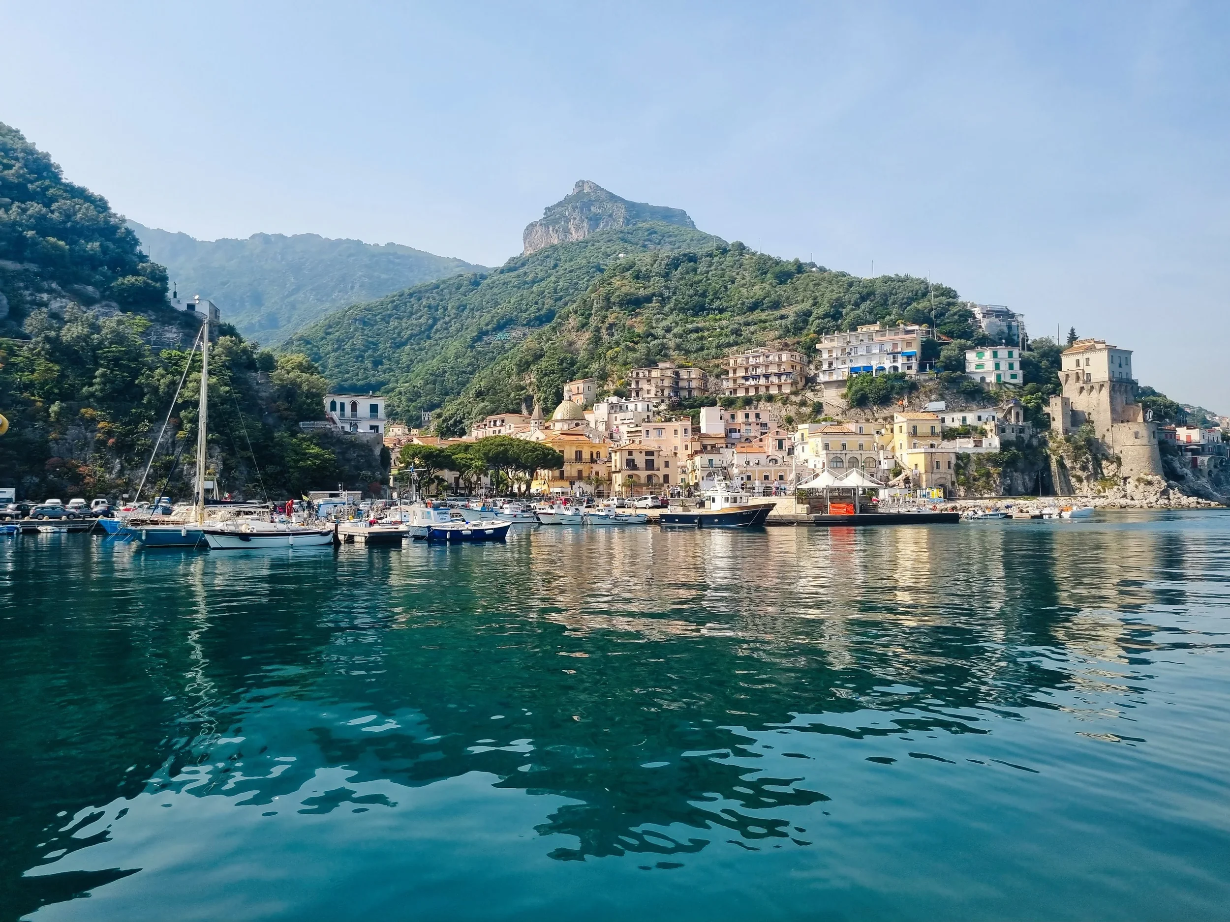 Cetara waterfront and coastal tower seen from the sea, with colourful houses beneath green hills.