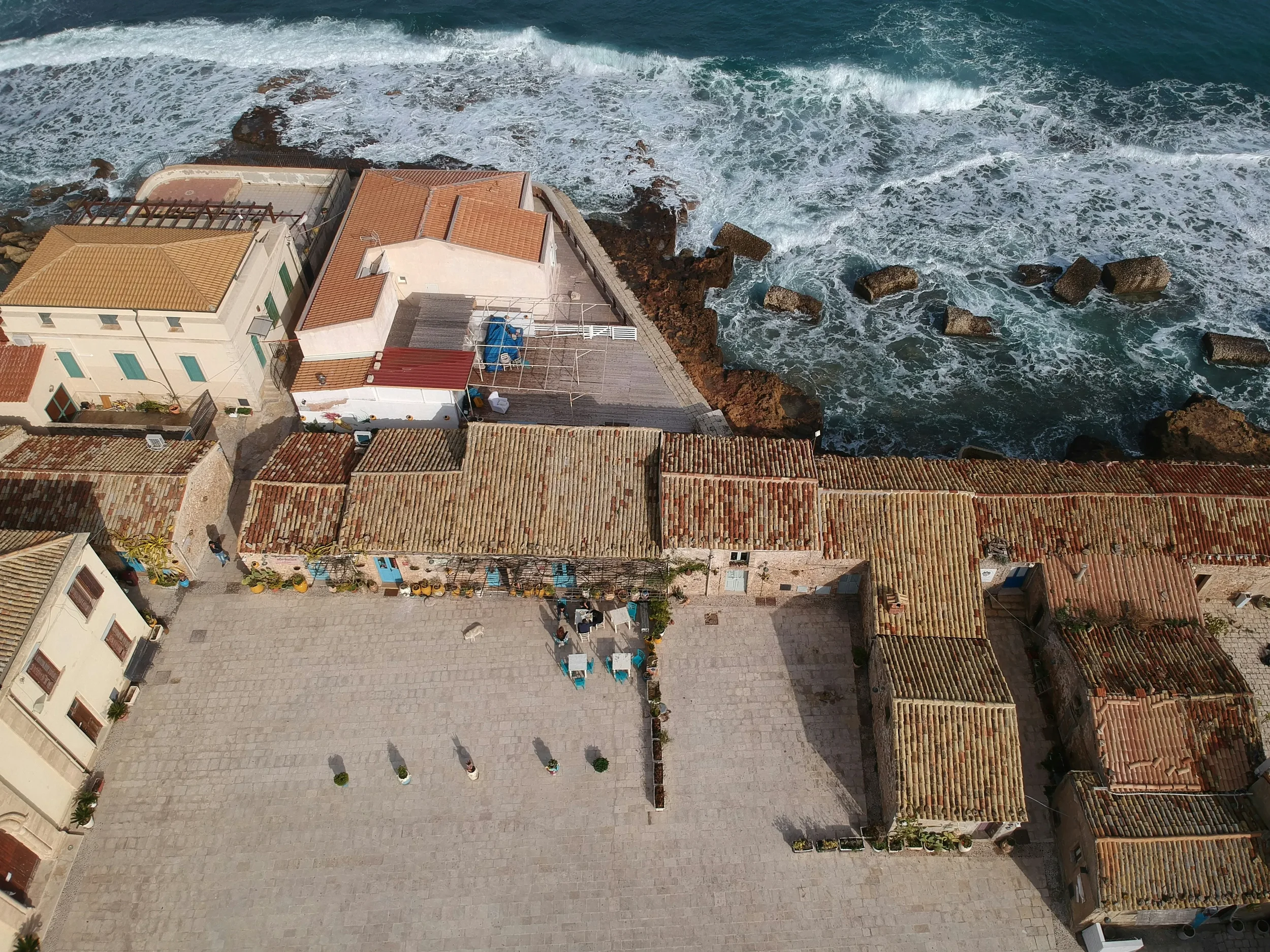 Aerial view of Piazza Regina Margherita and Marzamemi’s tiled waterfront roofs beside breaking waves