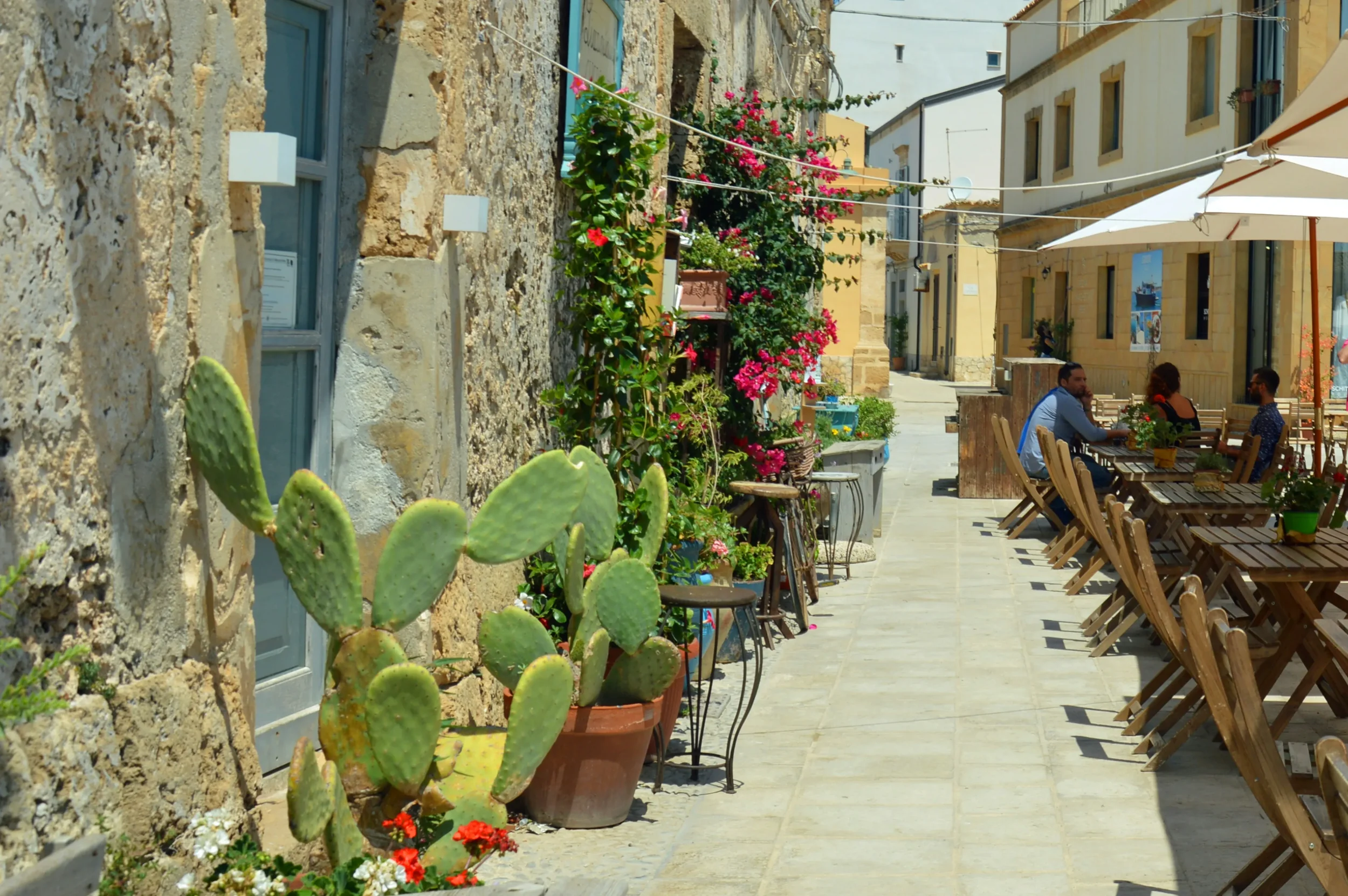 A sunlit lane in Marzamemi lined with stone walls, potted prickly pears and outdoor restaurant tables
