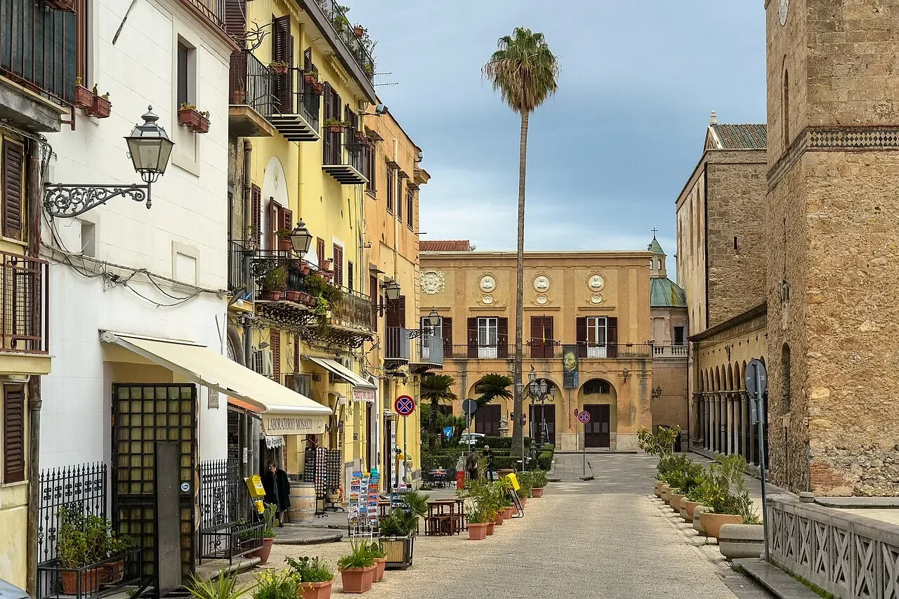 A pedestrian street beside Monreale Cathedral, with café awnings, balconies and a tall palm tree