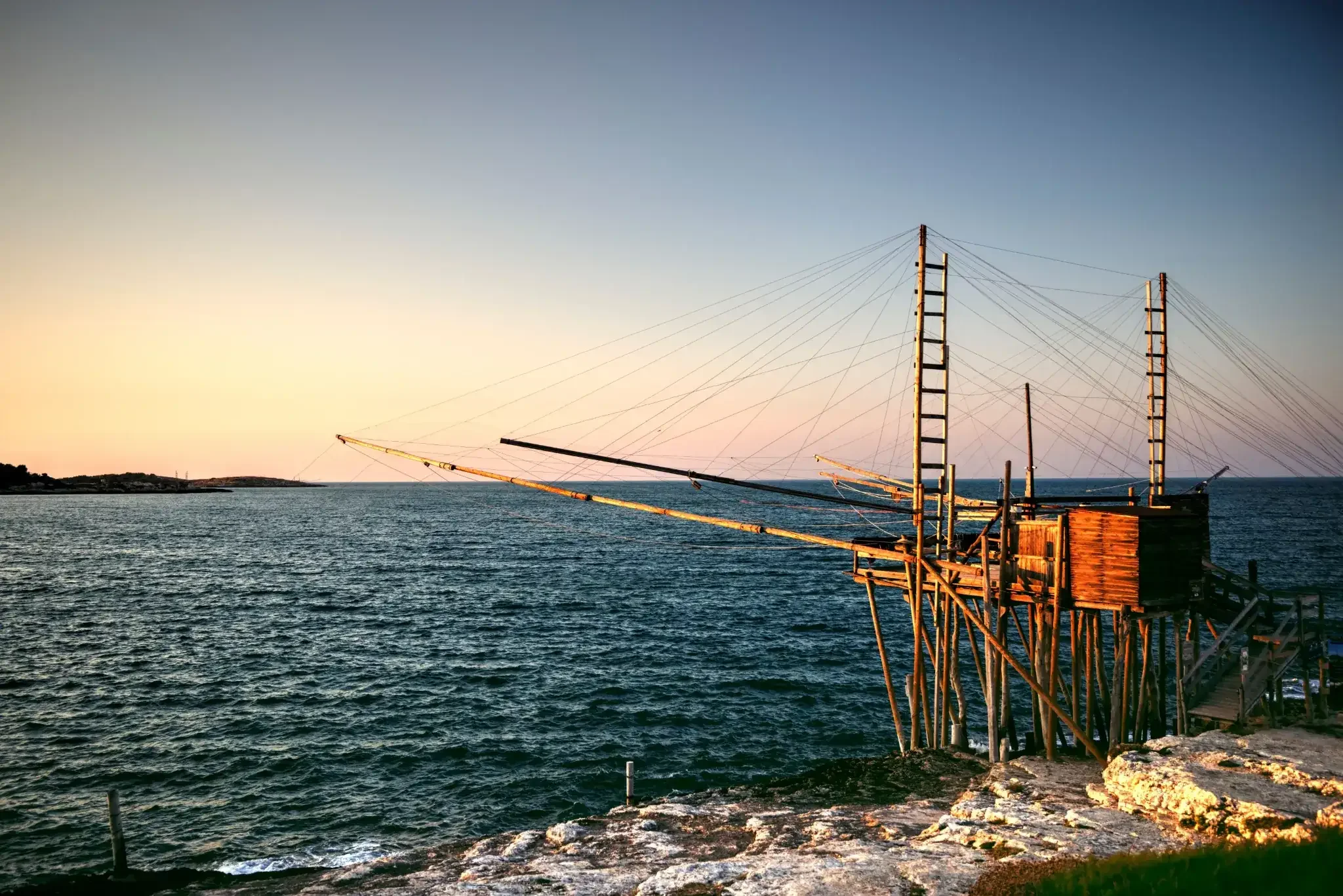 A coastal scene with a wooden pier and fishing equipment extending over calm water, under a sky transitioning from light to darker shades near sunset.