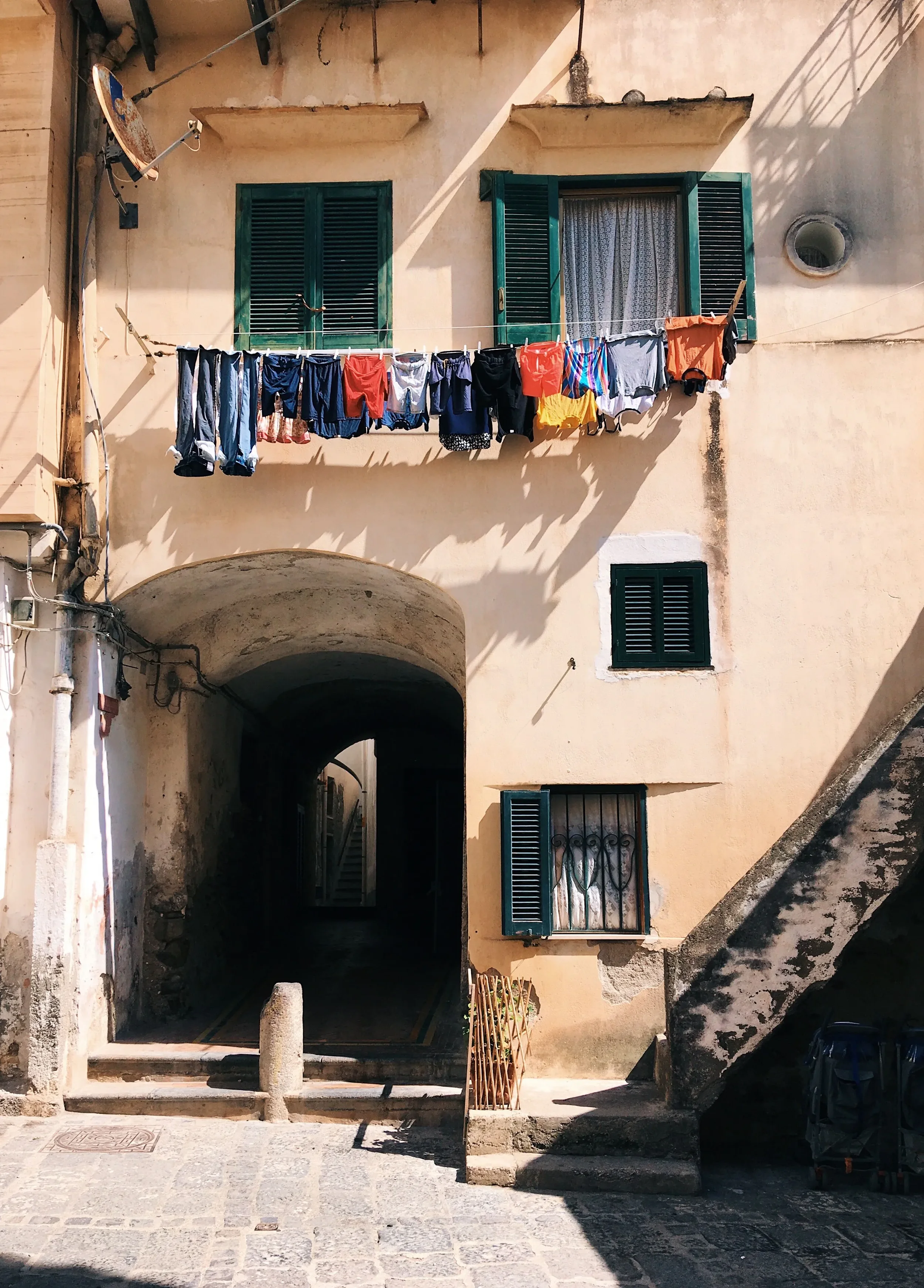Laundry hanging above an arched passageway, green shutters and stone steps in a sunlit village lane.