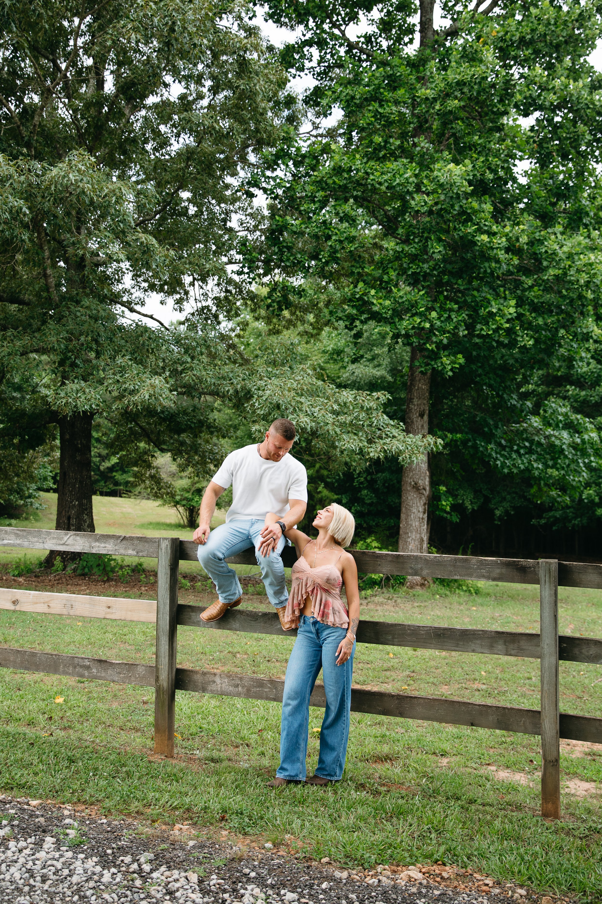 A man and a woman interacting outdoors near a wooden fence with trees in the background.
