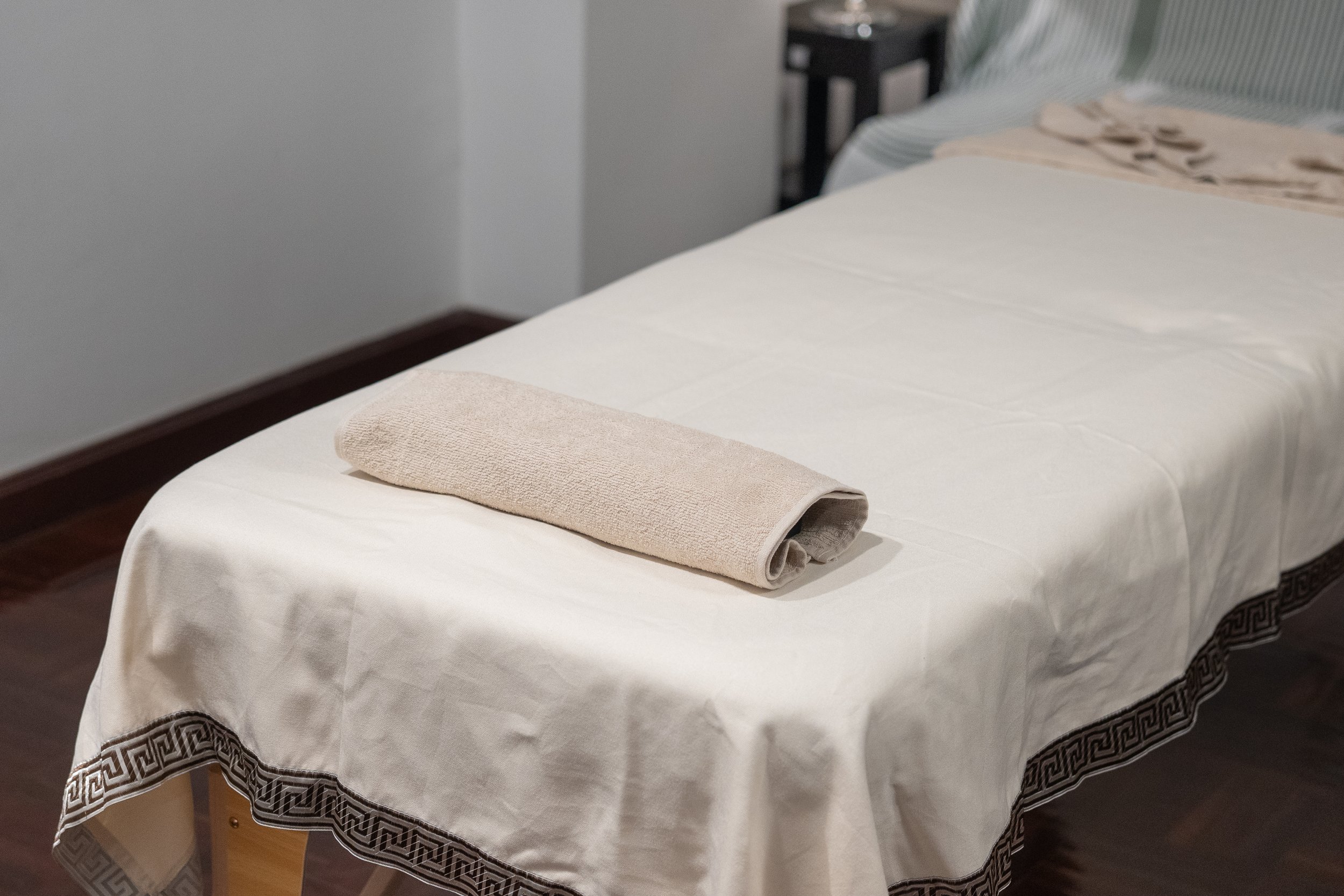 A clean massage table covered with a white sheet in a private room, with a rolled beige towel on top, in a room with wooden flooring and minimal decor.