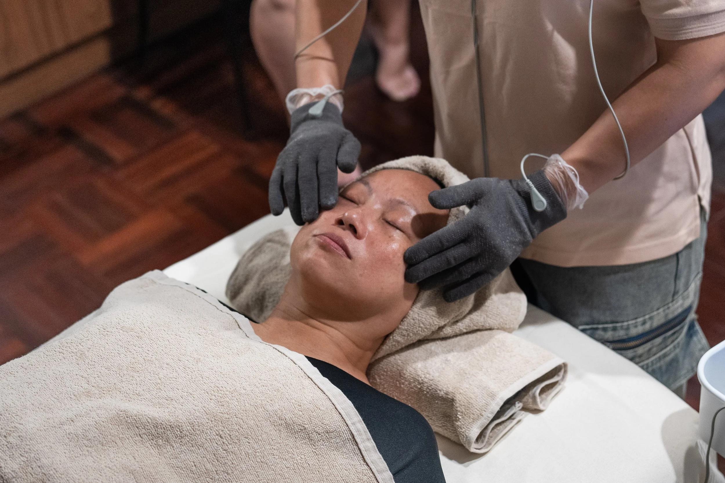 A lady receiving a microcurrent facial treatment from a practitioner in a spa to improve blood circulation and help reduce face puffiness