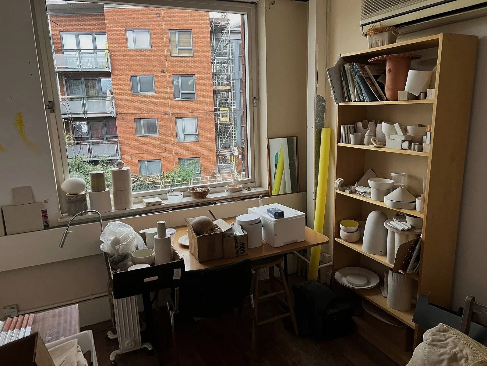 A cluttered room with a large window showing a brick building and fire escape outside. Inside, there is a wooden bookshelf filled with pottery and art supplies, a table with various items including boxes, jars, and a potato, and a black chair. The room appears to be used for art or pottery work.