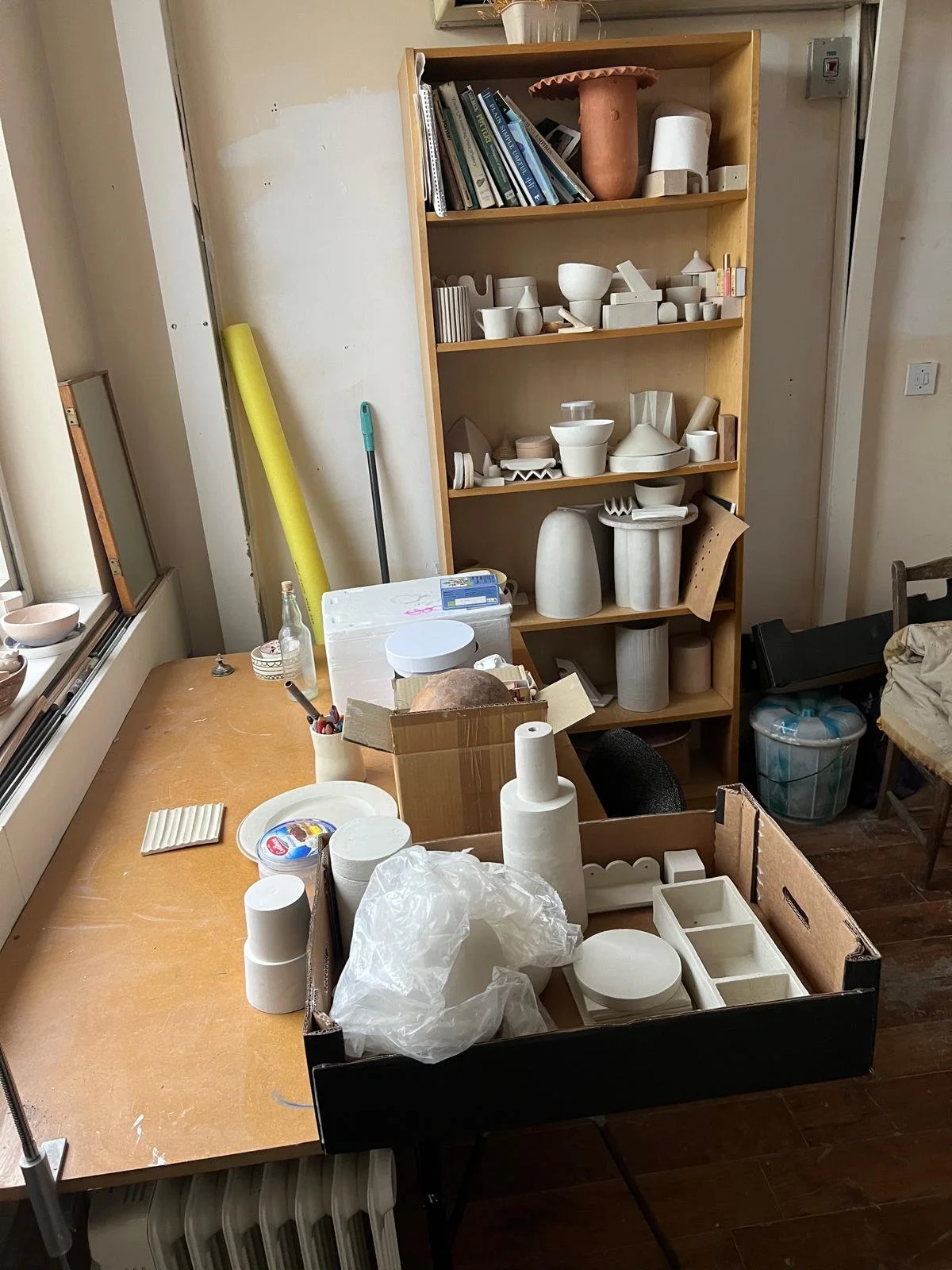 A pottery studio workspace with a wooden shelf filled with white ceramic pottery and books. A large table in front has various ceramic pieces, tools, and supplies for pottery making, with a box containing additional ceramic items.