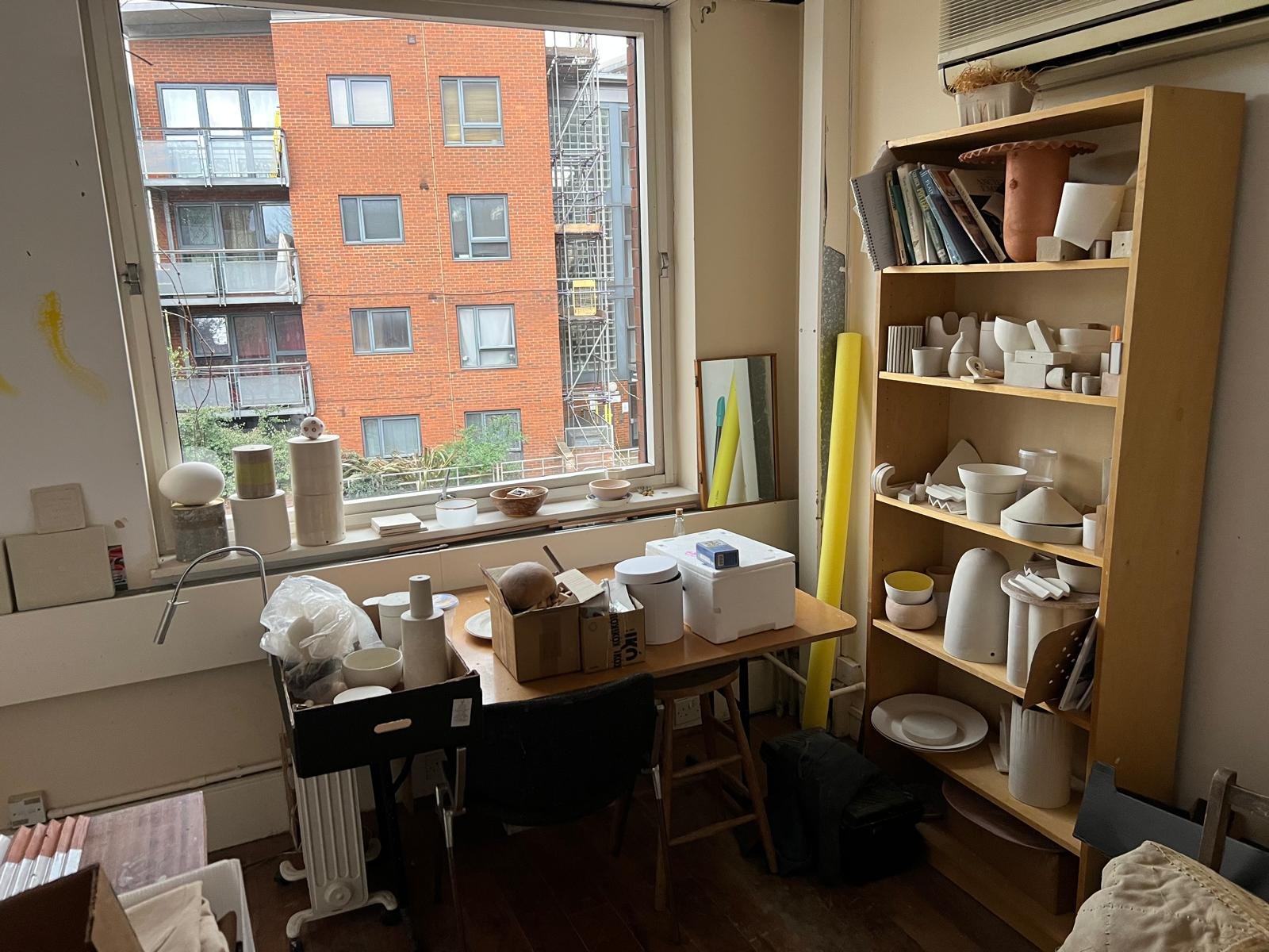A cluttered room with a large window overlooking a brick building, a wooden bookshelf filled with ceramics and art supplies, a table with boxes, and various art materials scattered around.
