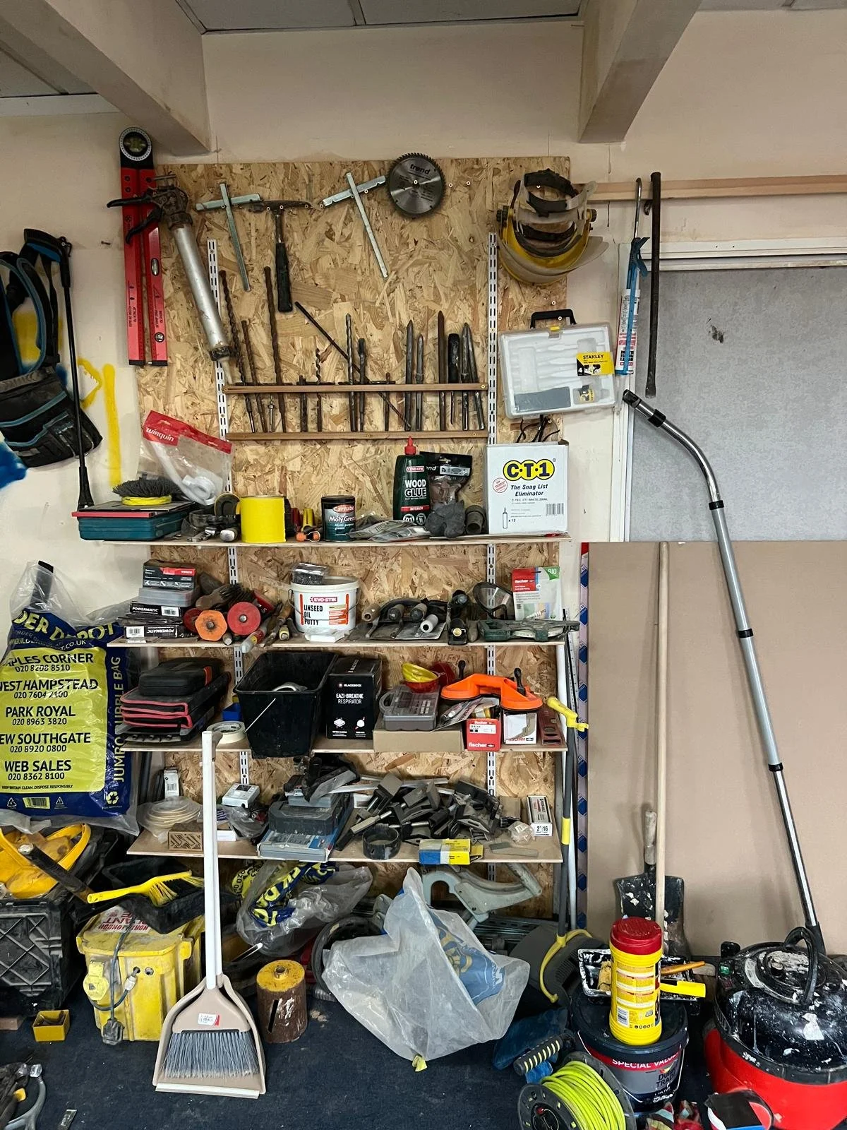 A garage wall with a large wooden shelving unit holding various tools and supplies, including drills, safety goggles, a saw, a level, and paint cans, among other items. The garage floor is cluttered with tools, a broom, a vacuum, and storage bags.