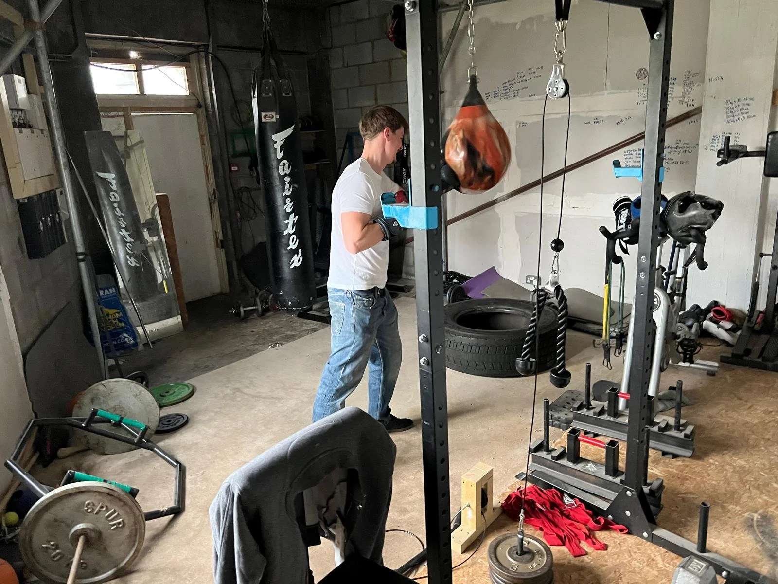 A young man wearing a white t-shirt, blue jeans, and boxing gloves is in a home gym, standing near a black power rack with various workout equipment around him, including weights, gloves, and punching bags.