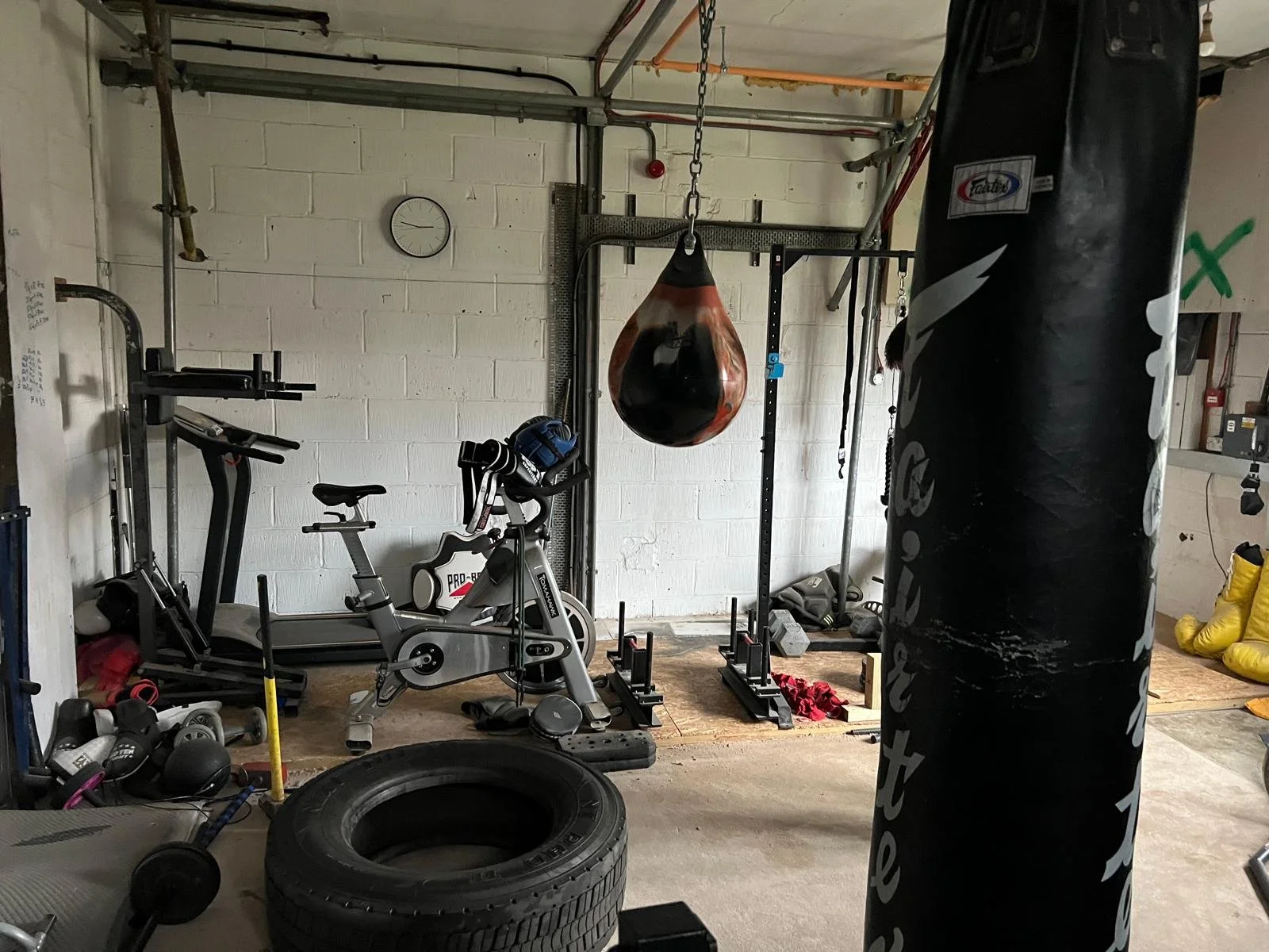 Home gym with exercise equipment including a treadmill, stationary bike, punching bag, and boxing gloves, all set up in a garage with a concrete floor and white brick walls.