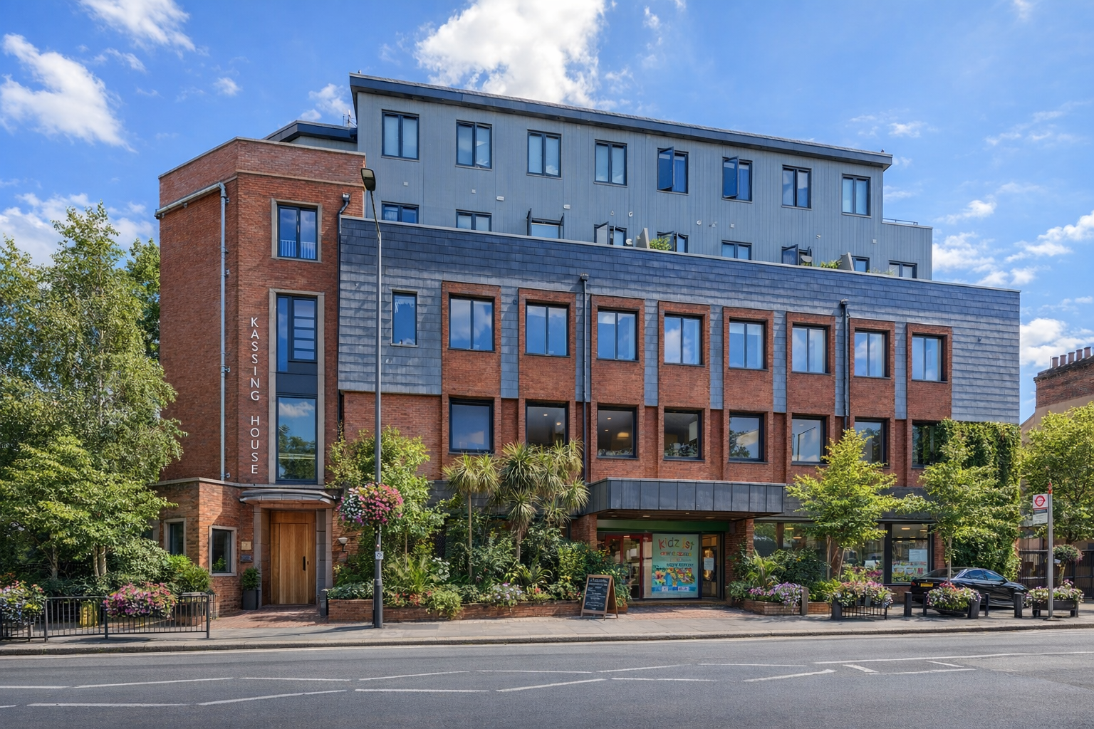 Multi-story building with brick and gray exterior, trees and flowers in front, a street, and a blue sky with clouds.