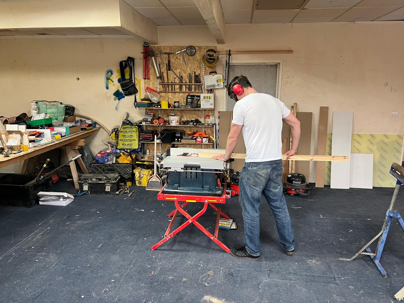 A man wearing a white t-shirt, jeans, and red ear protection uses a miter saw in a cluttered woodworking workshop.