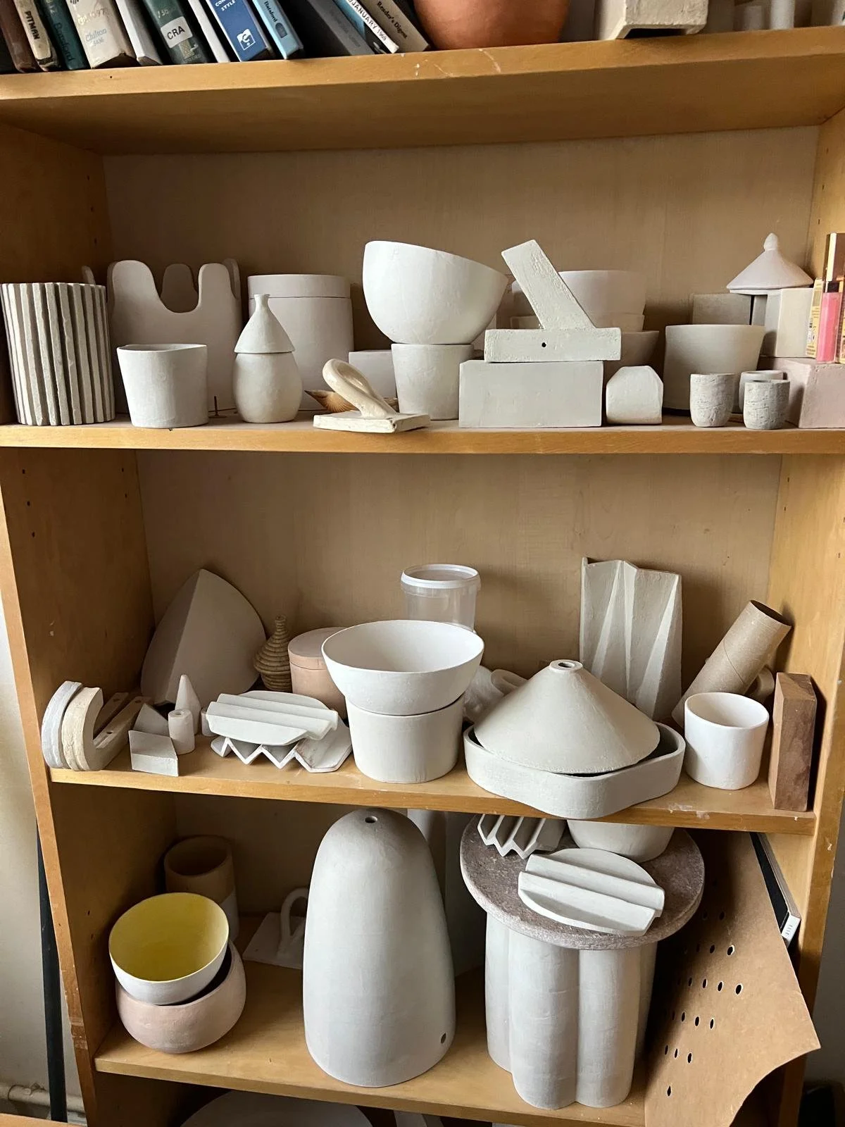 Wooden bookshelf filled with unfinished ceramic pottery and sculpting pieces, including bowls, vases, and abstract forms.