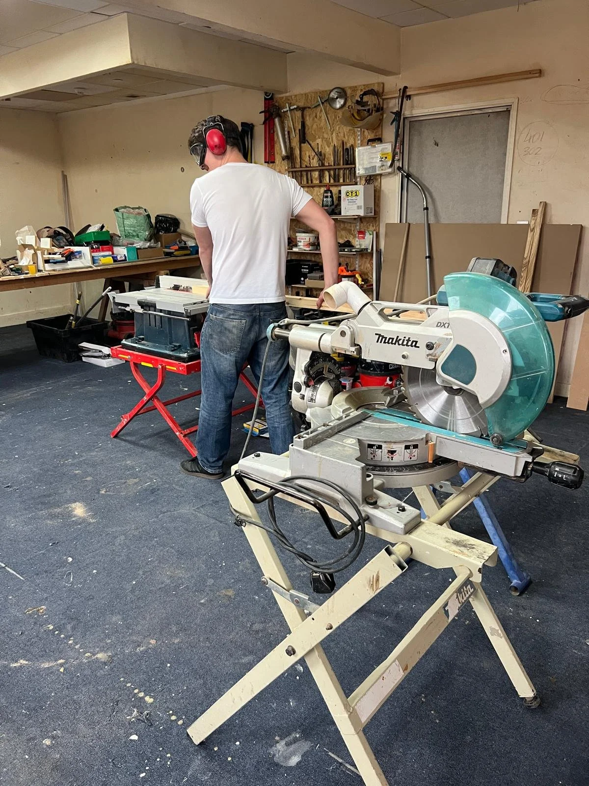 A person wearing a white t-shirt, jeans, and safety earmuffs working in a woodworking shop with a large Makita chop saw in the foreground. The workshop has tools and supplies on workbenches and shelves.