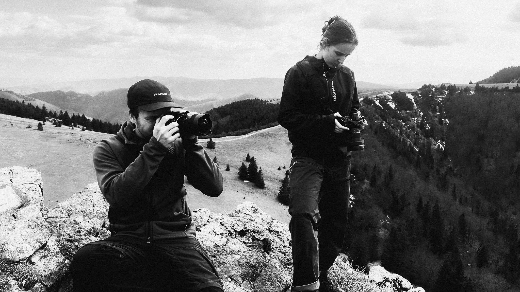 Two photographers on a rocky mountaintop, one sitting and looking through a camera, the other standing and examining a camera, with a scenic landscape of hills, trees, and snow patches in the background.