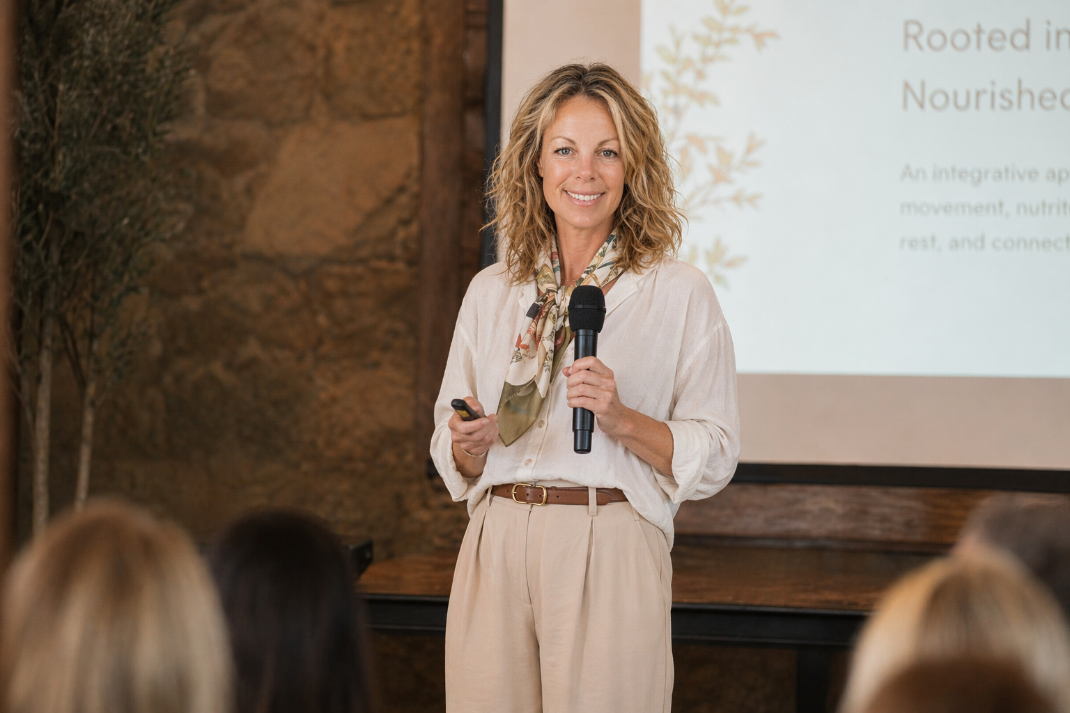 A woman with curly blonde hair giving a presentation in front of an audience. She is holding a microphone and a remote control, standing near a projection screen with text.