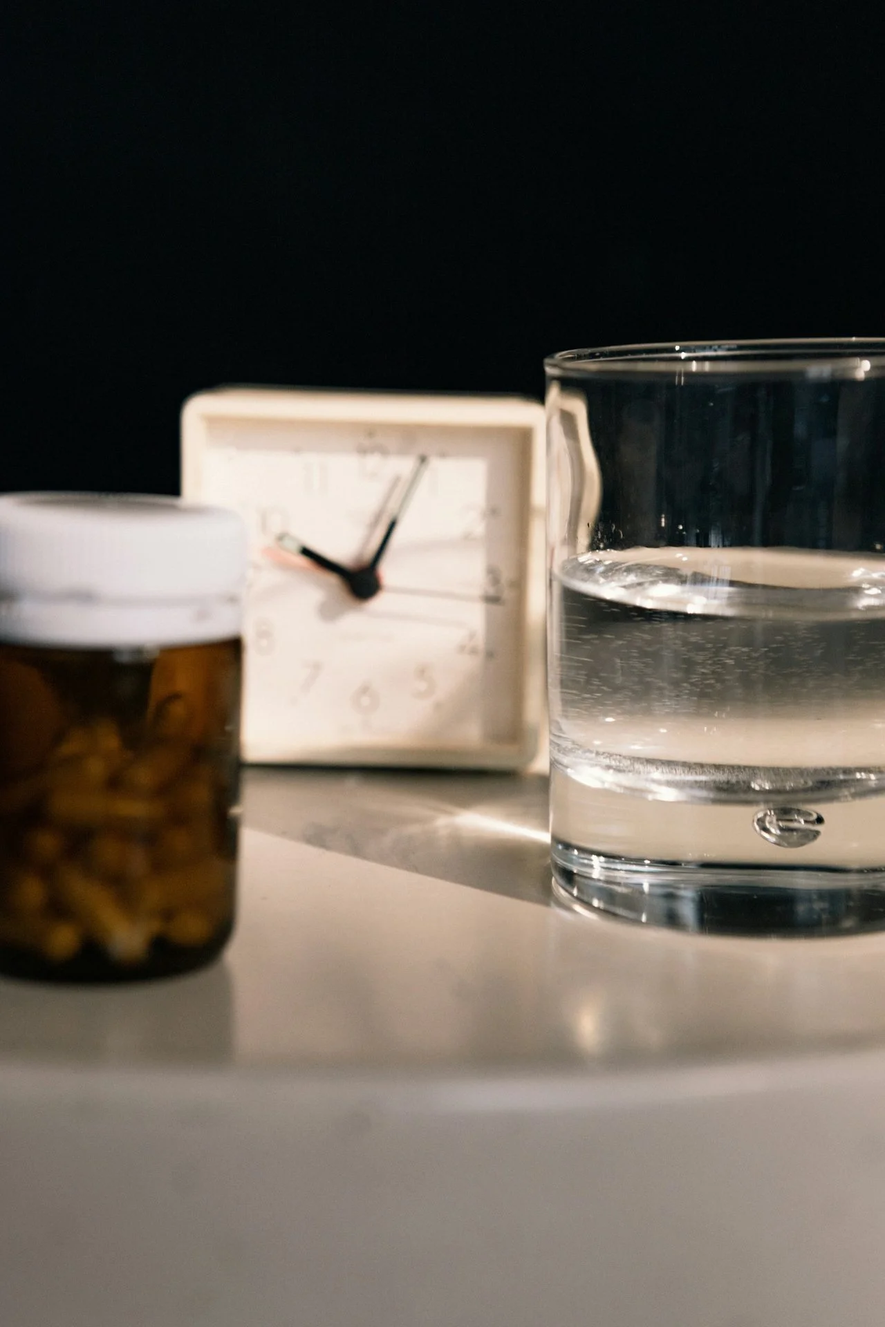 A glass of water, a prescription pill bottle, a small white clock, and an empty transparent cup on a white surface against a black background.